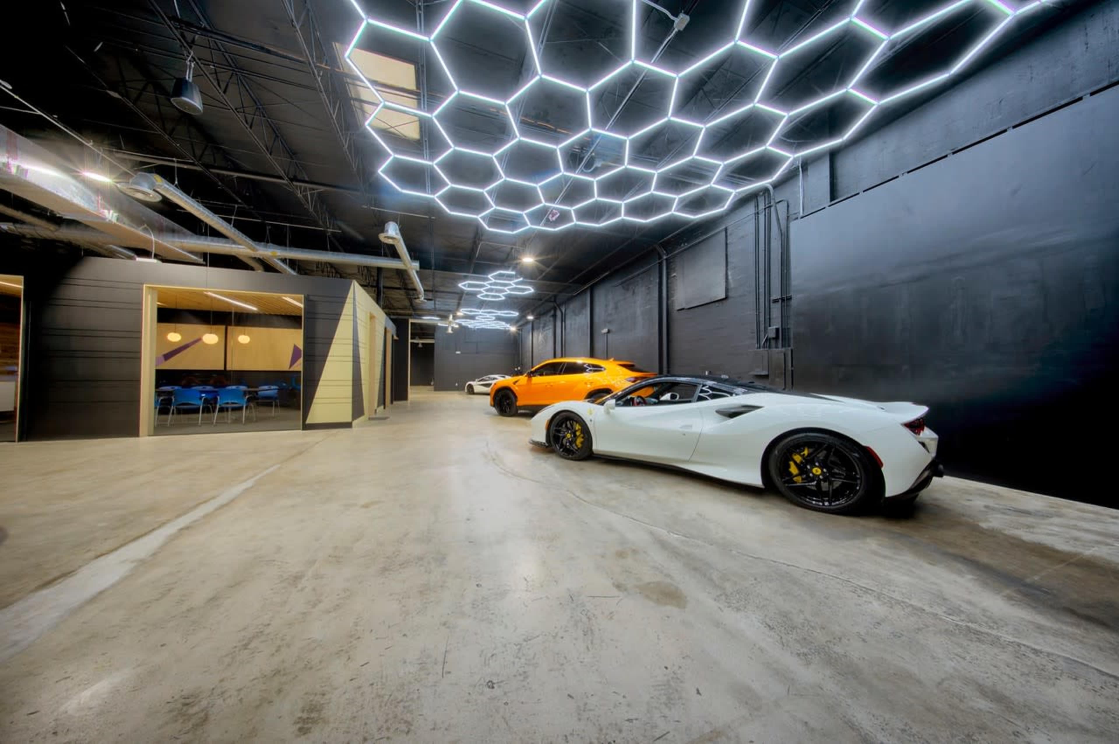 A spacious, modern garage features a white sports car and an orange vehicle under a hexagonal light installation, with a concrete floor and black walls.
