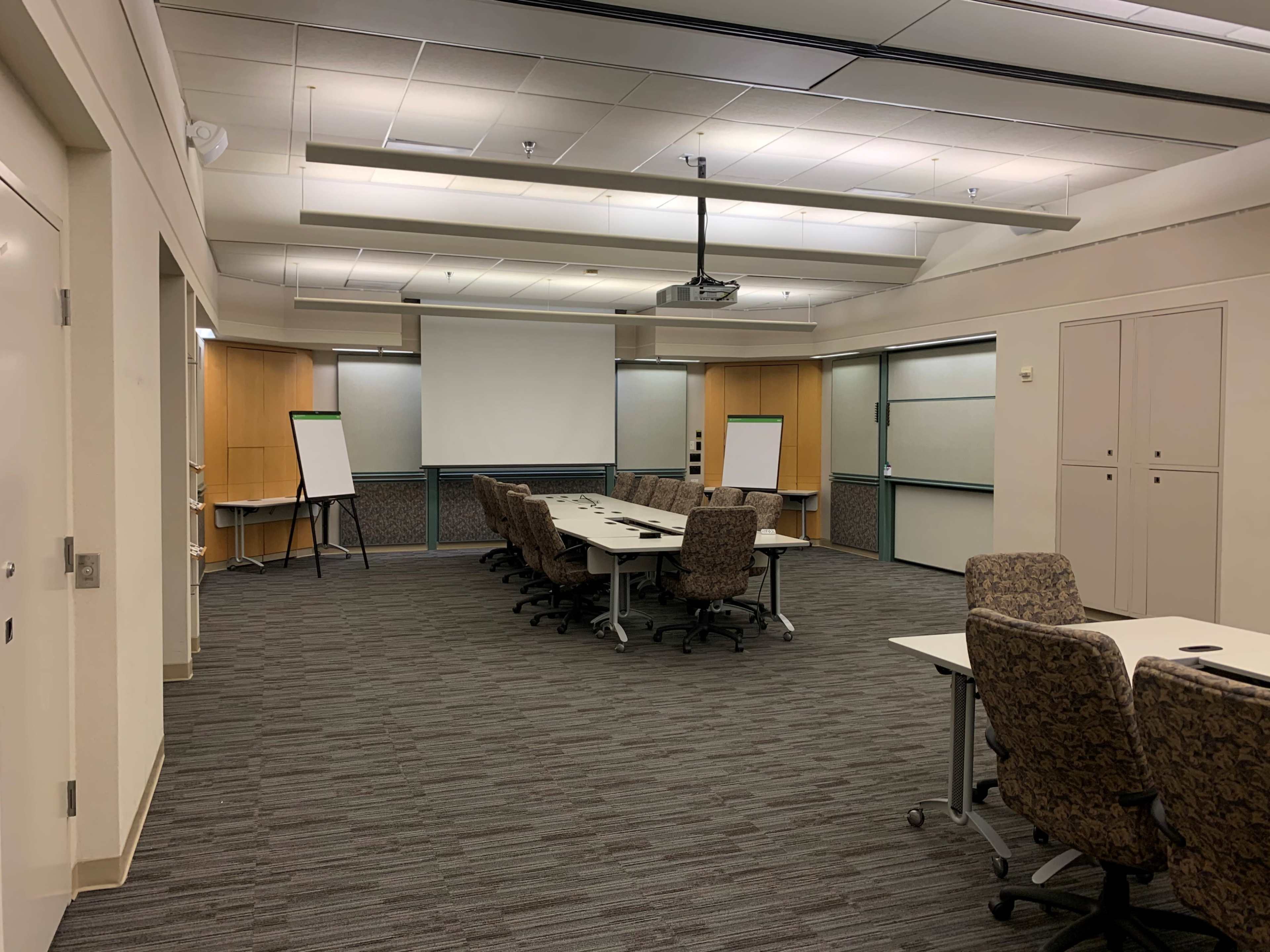A conference room with a rectangular table surrounded by chairs, a projector screen at the front, and flip charts positioned at each end.
