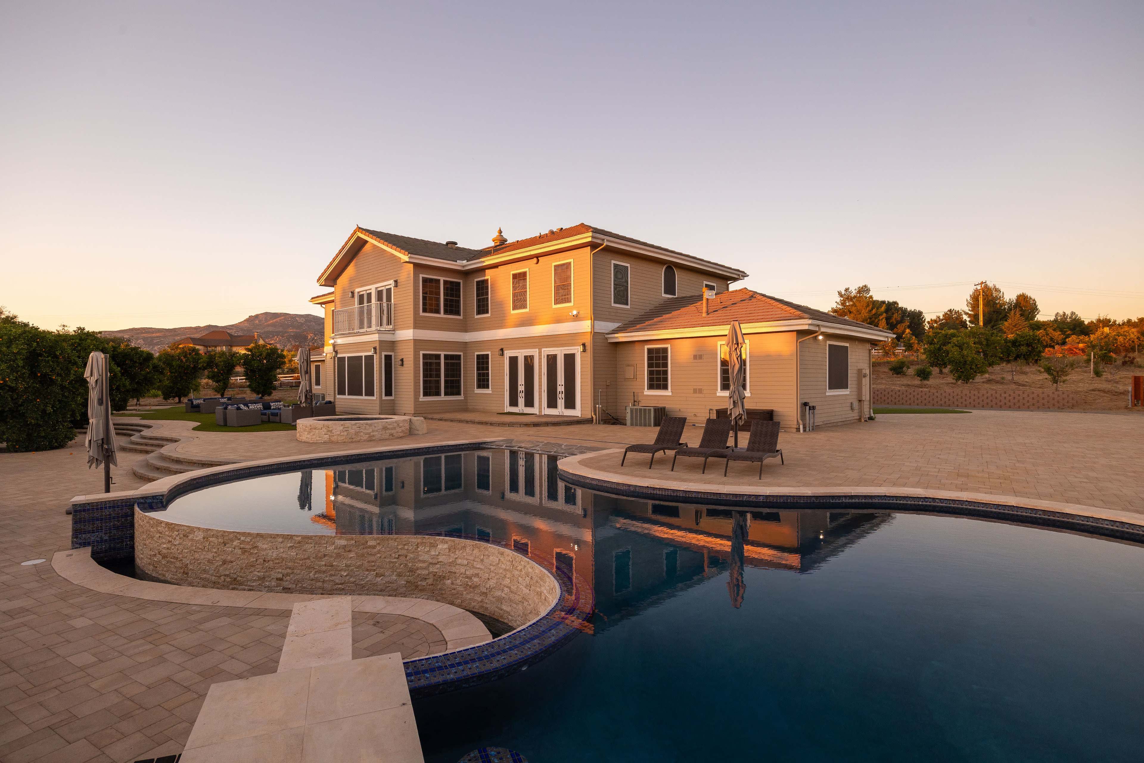 A large two-story house with a swimming pool and patio is set against a backdrop of hills at sunset.
