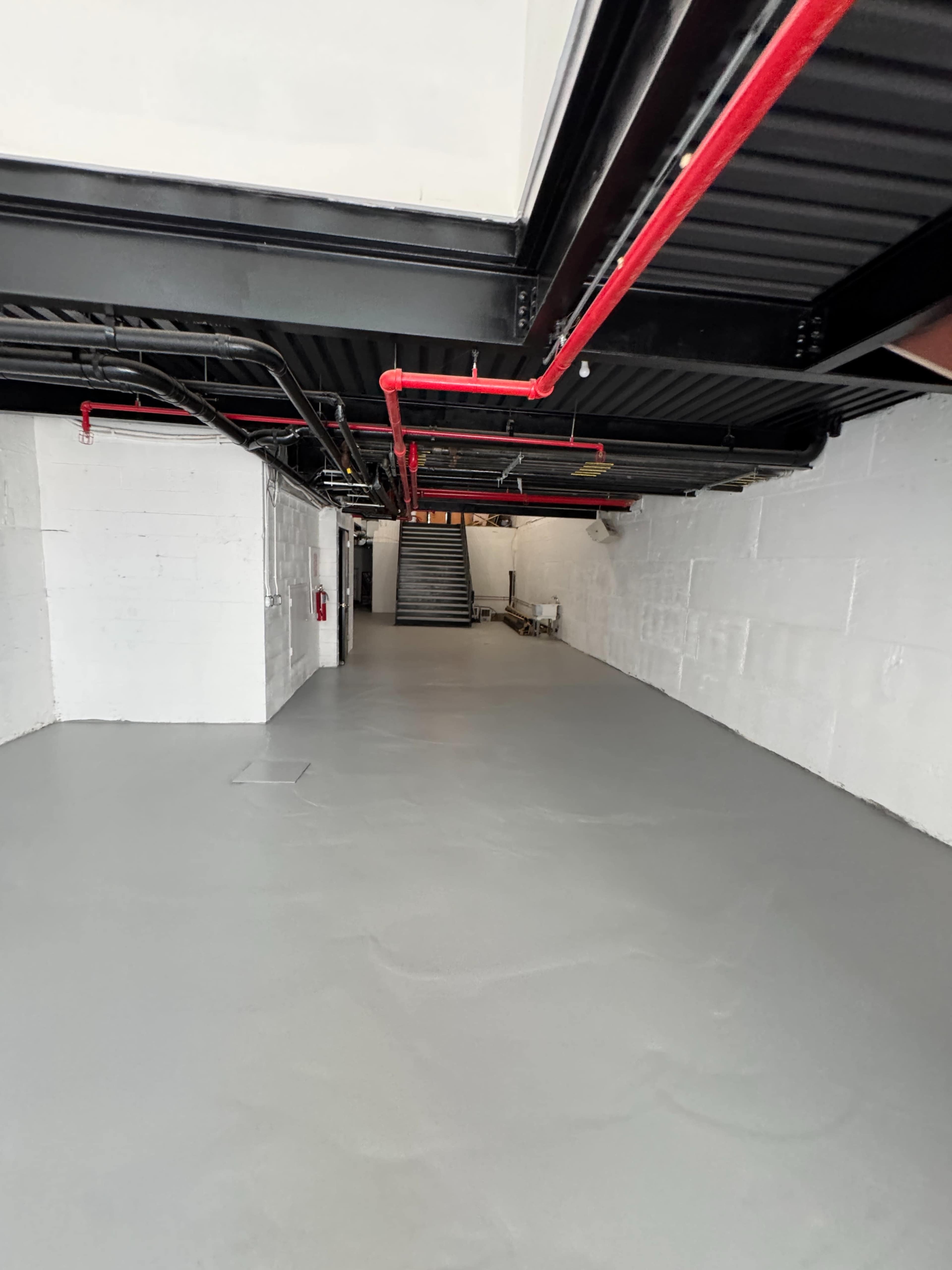 The image shows a spacious, unfinished basement with gray flooring, exposed black beams, and red pipes running along the ceiling.