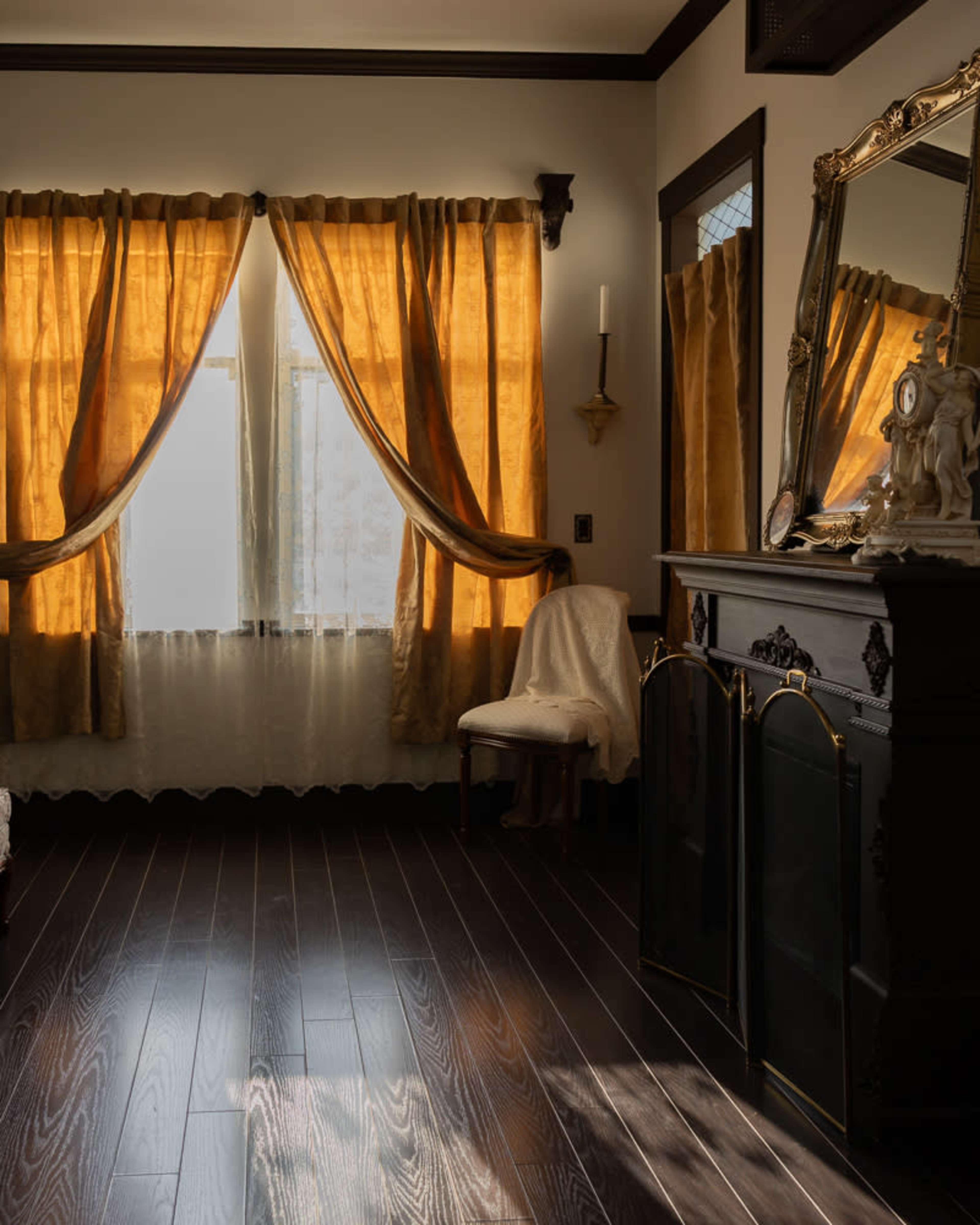 The room features large windows draped with gold curtains, a mirror above a dark wooden dresser, and a chair with a white covering against the wall.