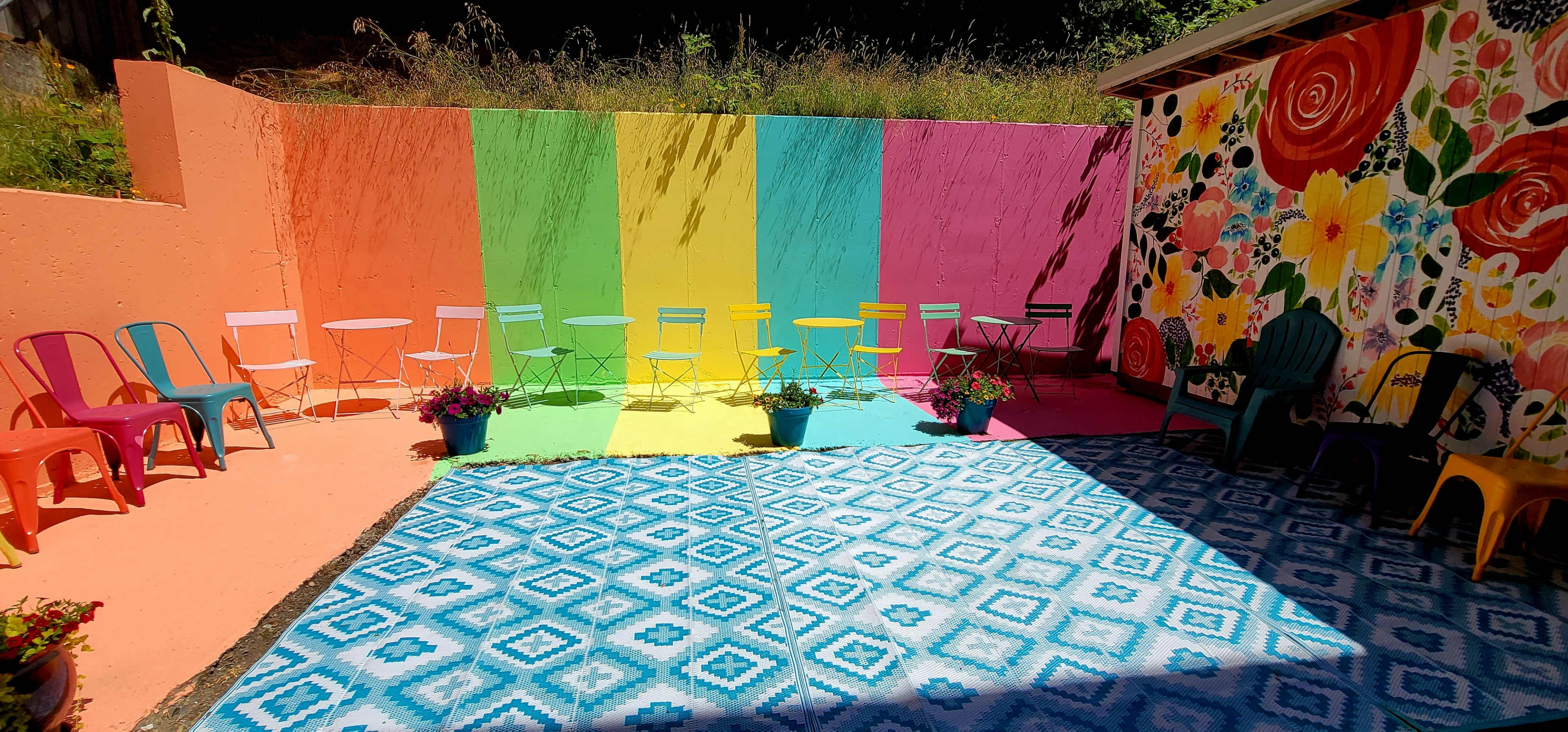 A vibrant outdoor space features colorful seating arranged along a striped mural, with a patterned rug and flowering plants enhancing the scene.