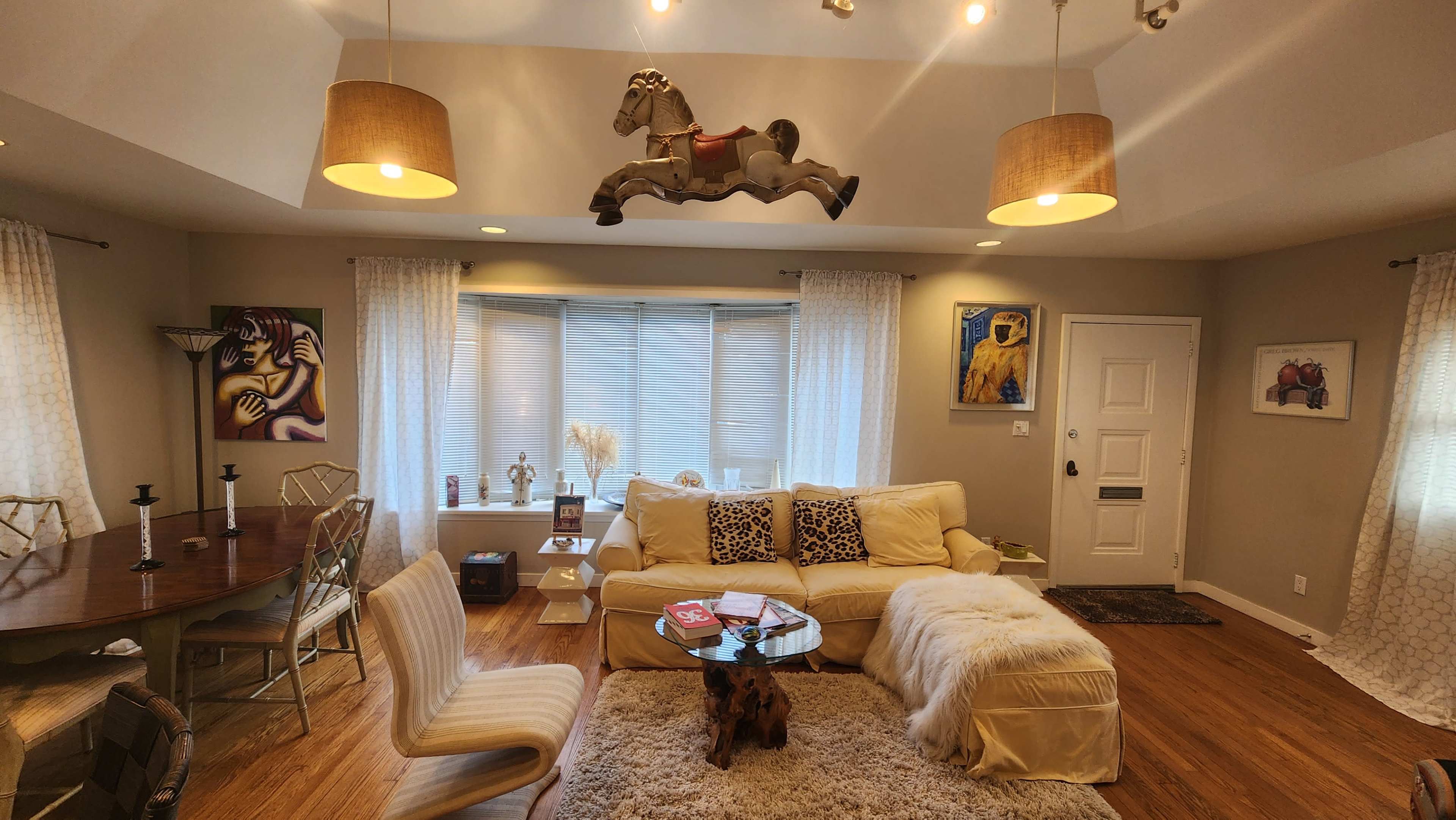 A spacious living room features a cream-colored sectional sofa, a wooden dining table, and a mounted horse sculpture above a window displaying sheer curtains.