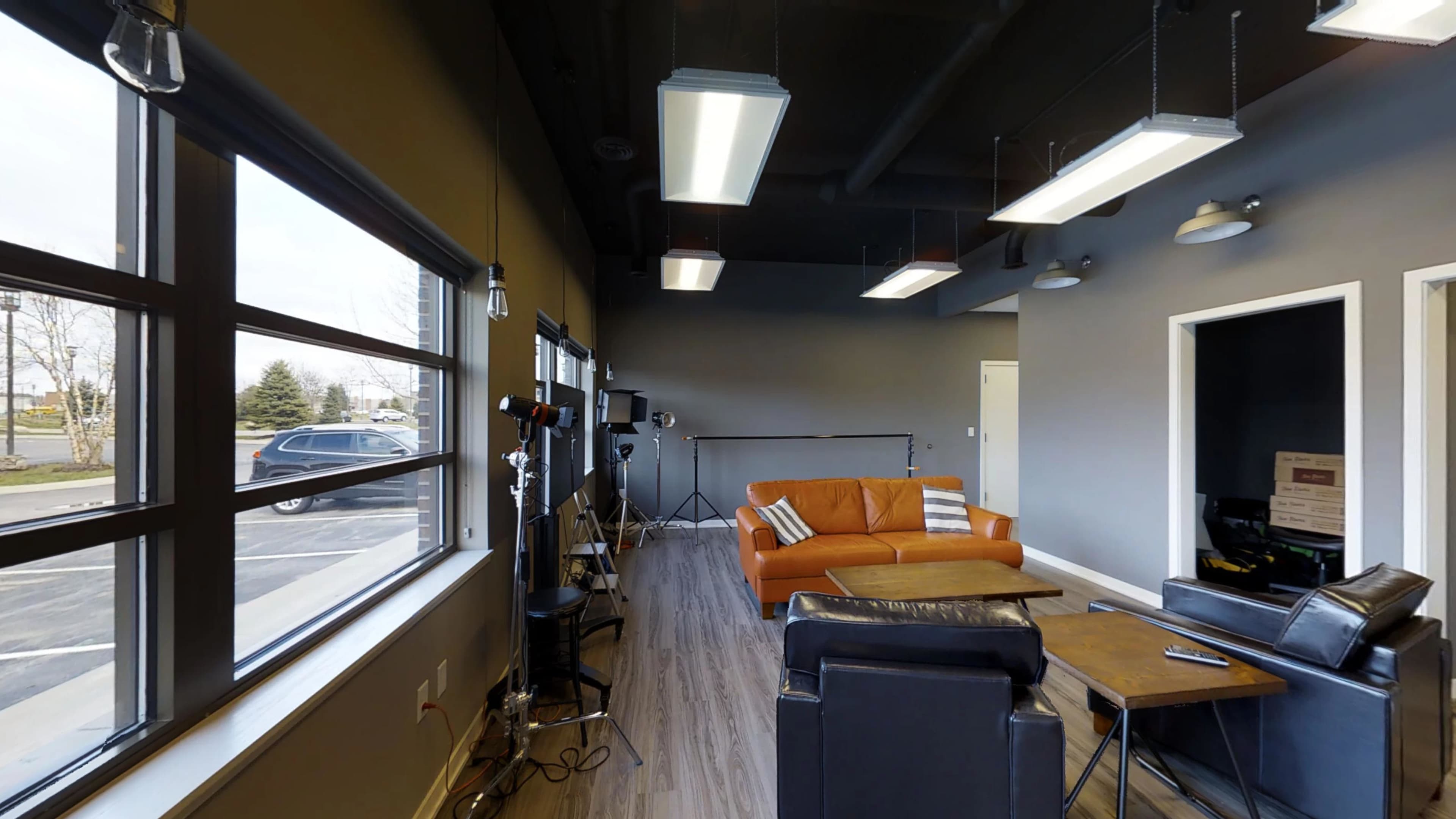 A modern interior of a lounge area featuring a brown sofa, a black armchair, and studio lighting equipment.
