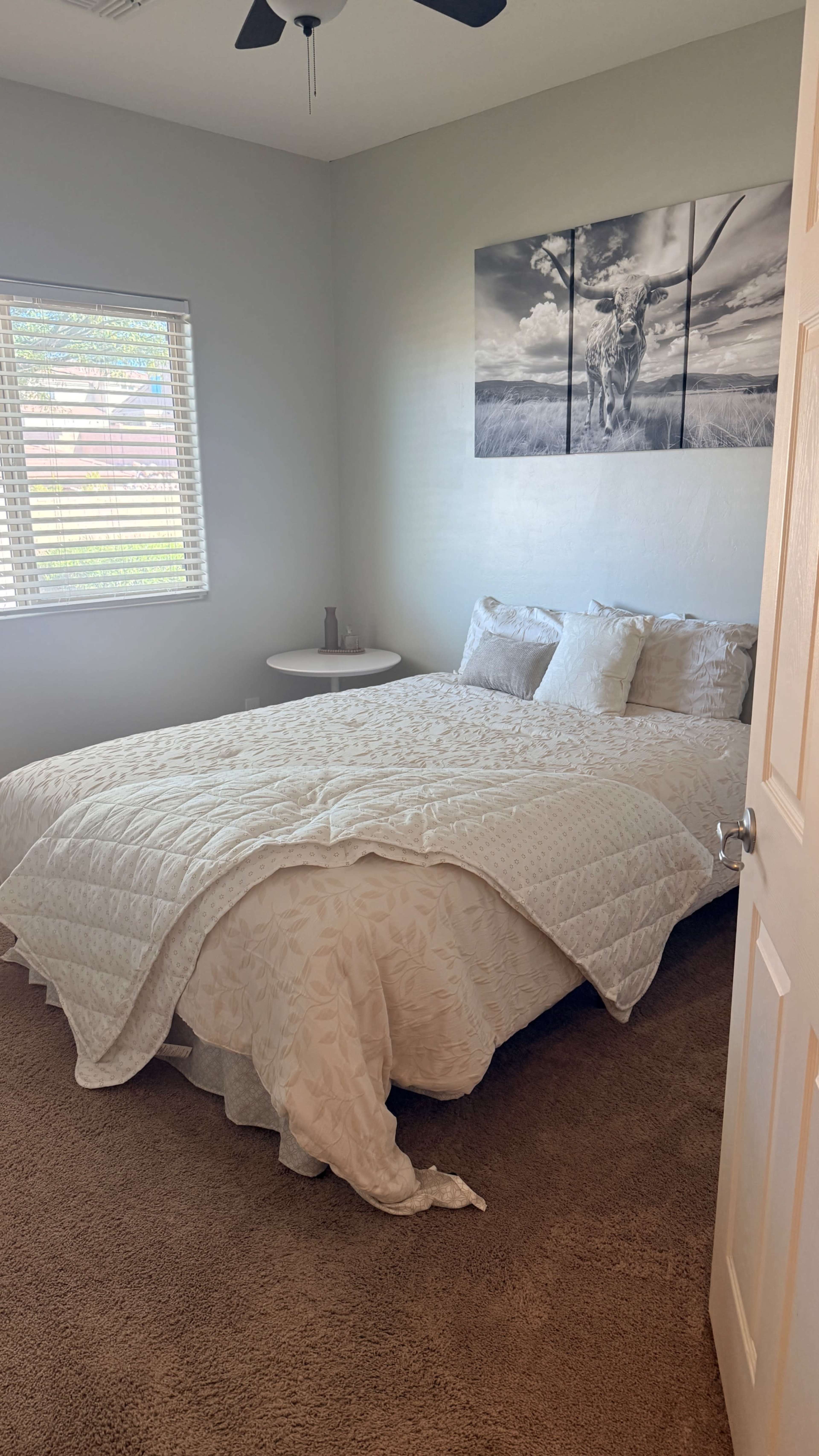 The image shows a neatly arranged bedroom with a large bed covered by a white quilt, a small round table beside it, and a wall featuring a black-and-white photograph.