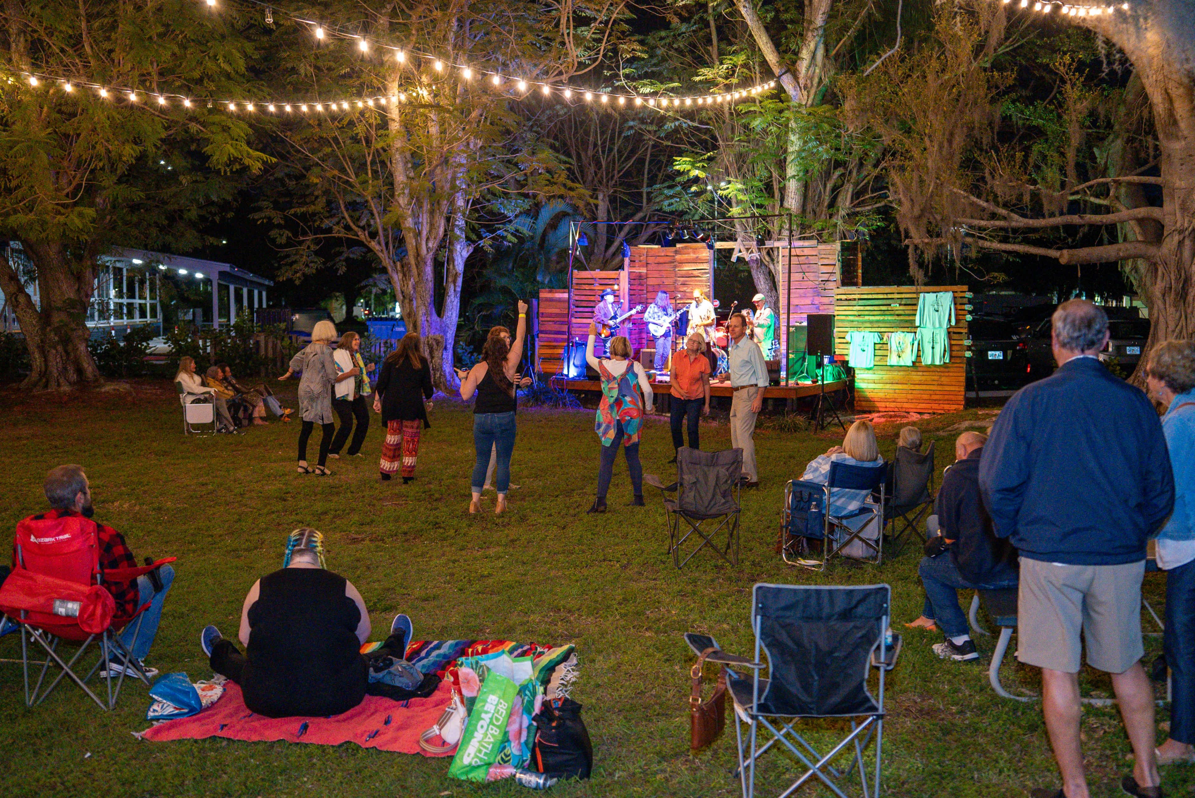 A group of people watches a live band perform on a stage illuminated by string lights in a park at night.