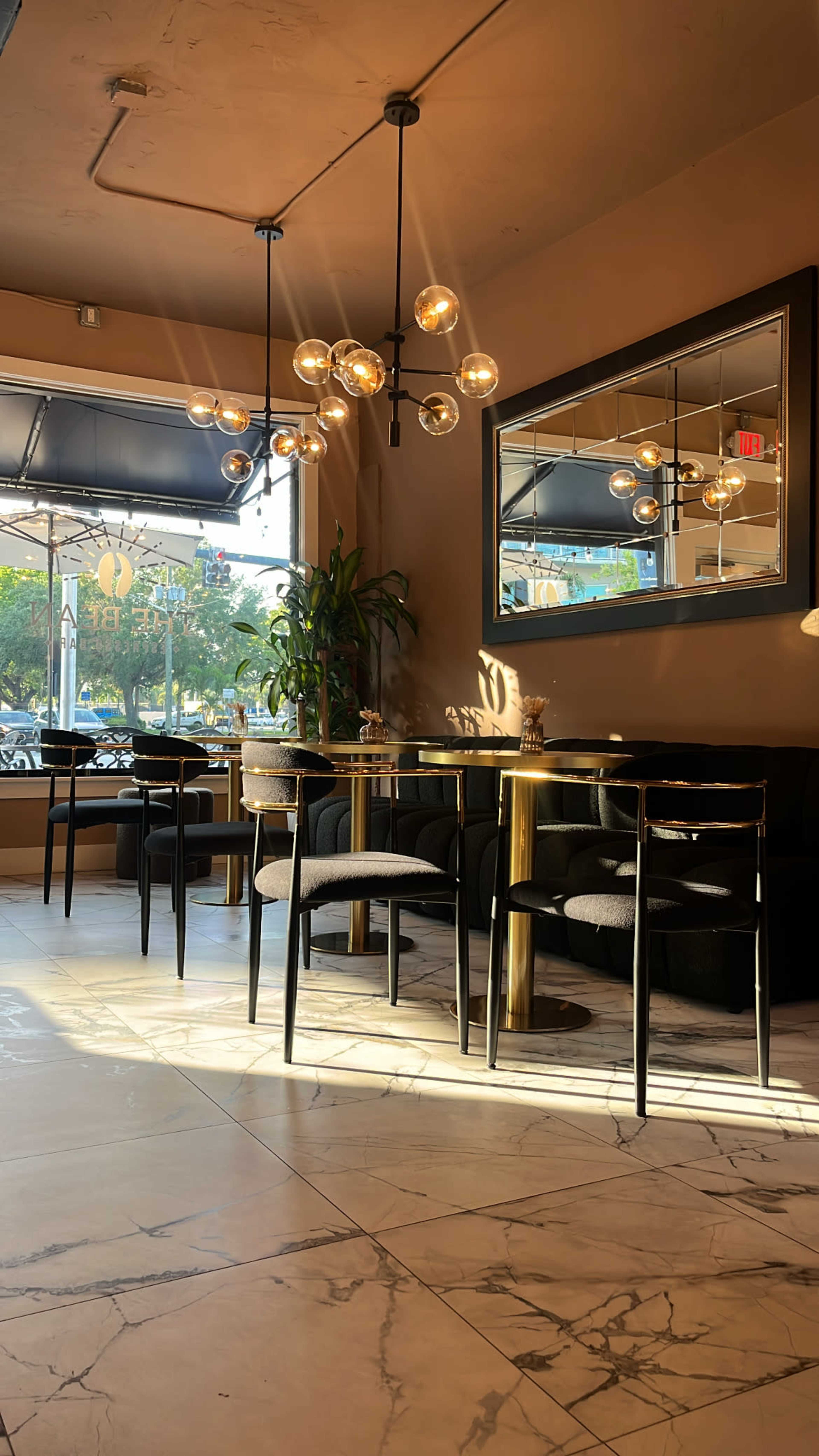 The image shows a modern cafe interior with a mix of round tables and black seating, illuminated by natural light coming through large windows and featuring a decorative chandelier and a wall mirror.