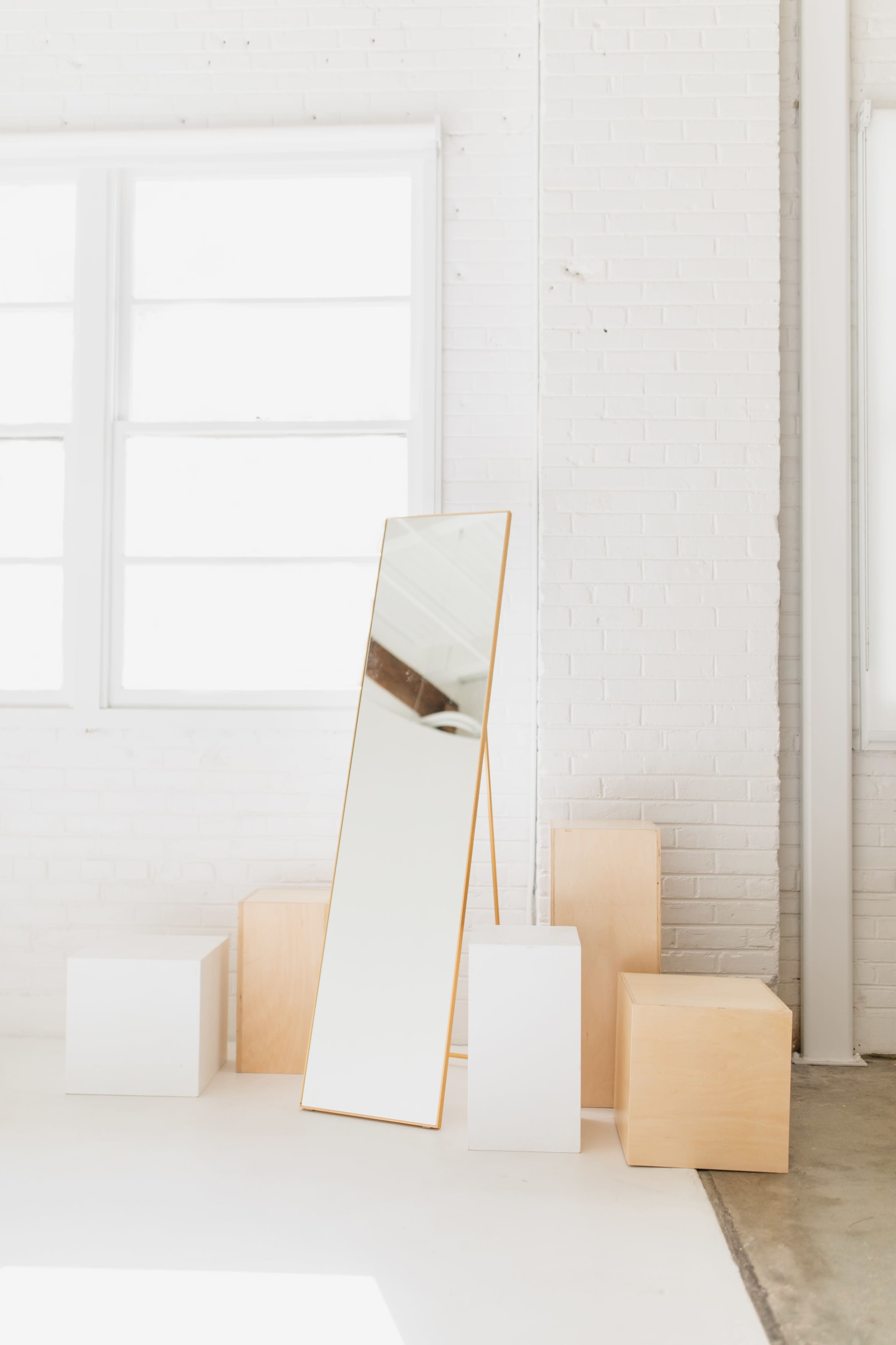 A tall mirror with a wooden frame leans against a white brick wall near several geometric wooden boxes.