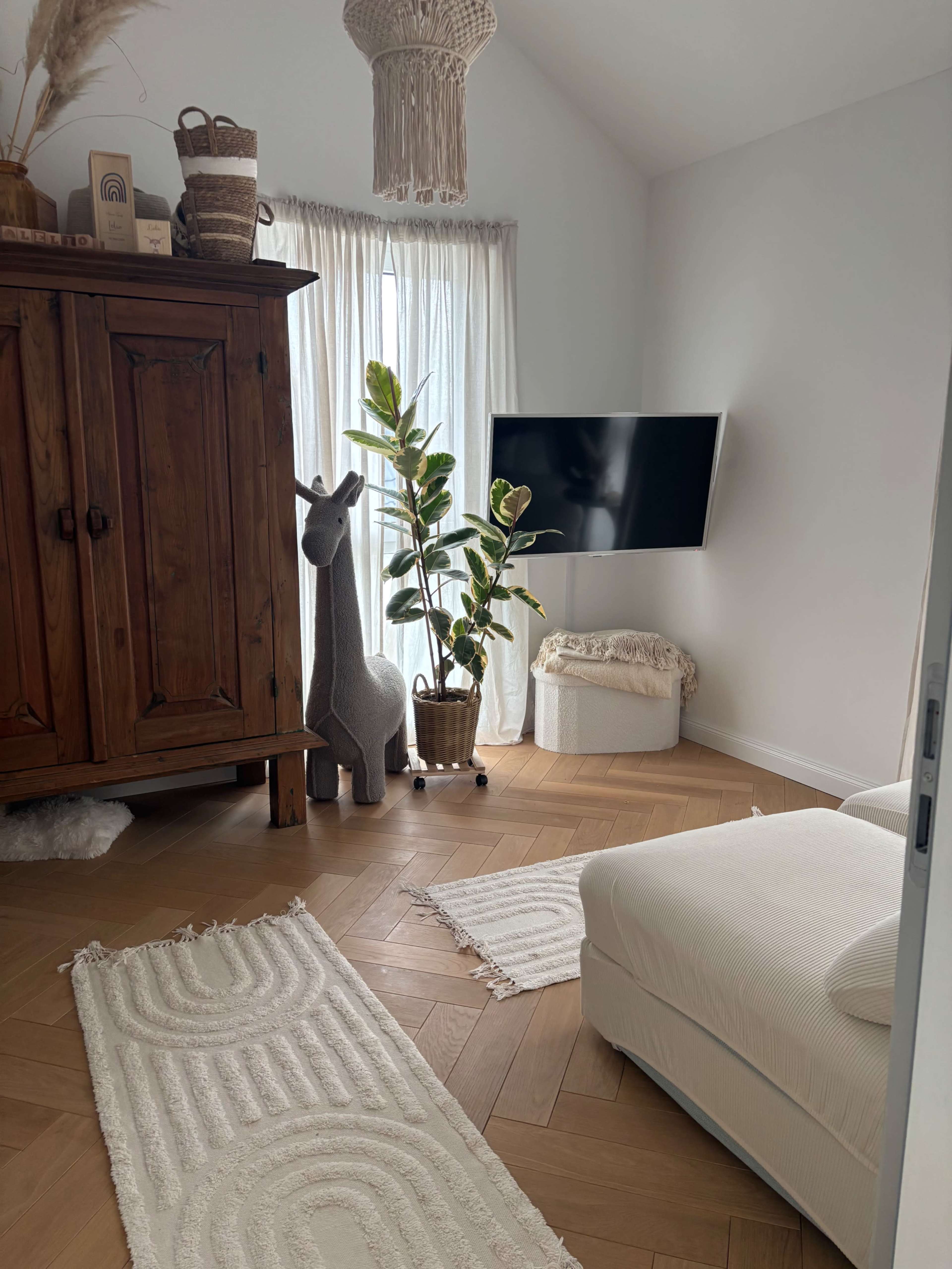The image depicts a cozy room with a wooden wardrobe, a tall indoor plant, a television mounted on the wall, and light-colored rugs on a herringbone floor.