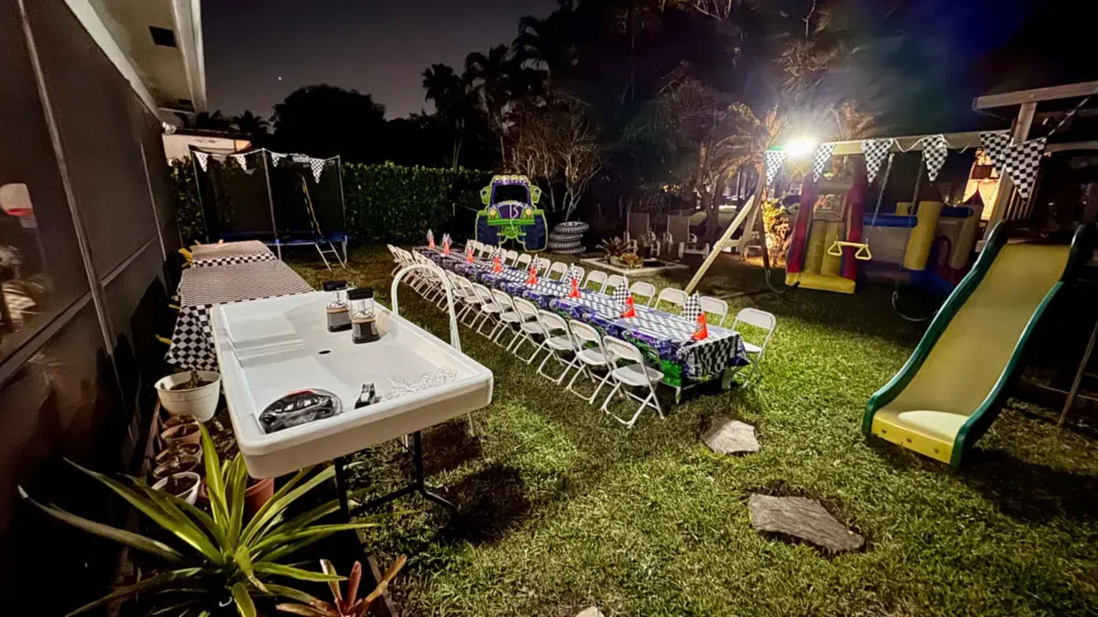 The backyard features a long table set with chairs, a playground structure, and various decorations under dim lighting.
