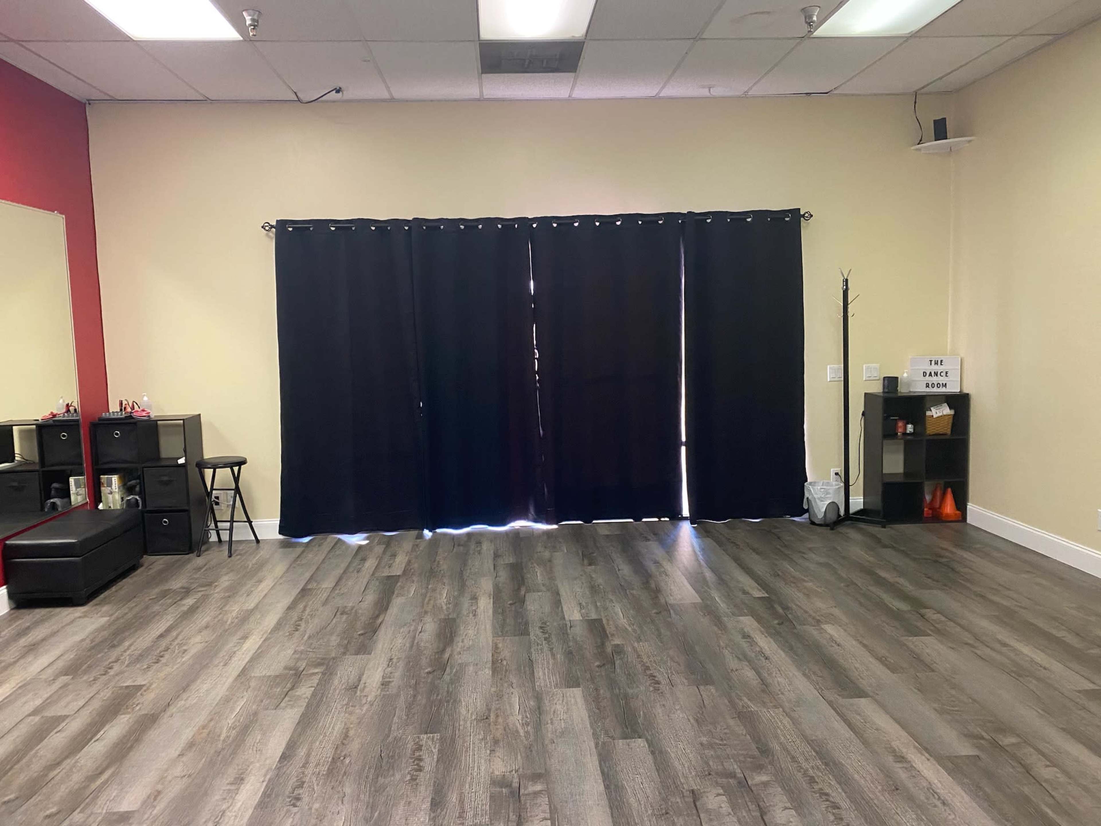 The image shows an empty room with a wooden floor, a large black curtain covering a window, and minimal furnishings including a chair, a small table, and a shelf against one wall.