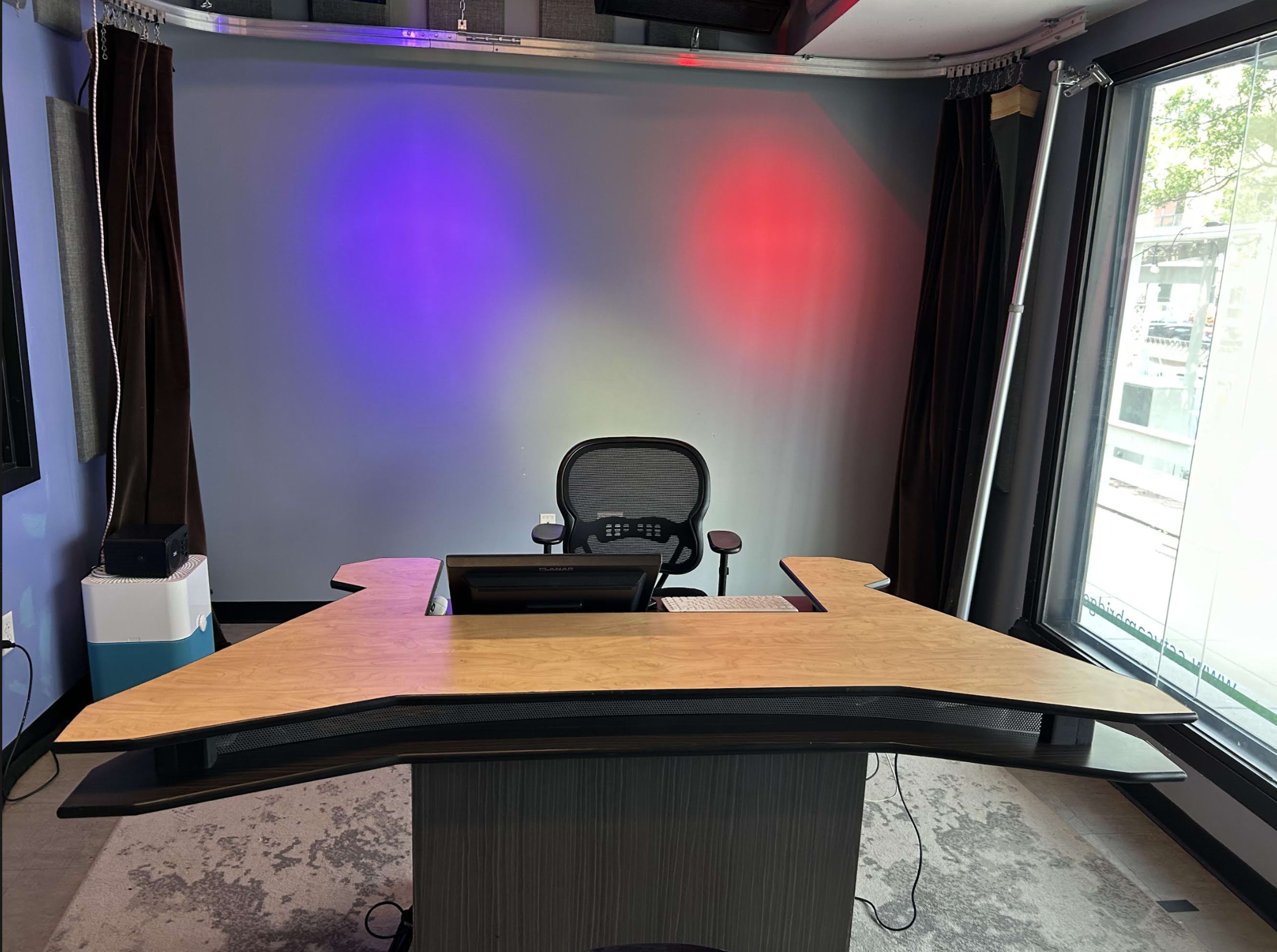 The image shows a modern desk with a unique angular design, positioned in a room with colorful lighting and a large window.