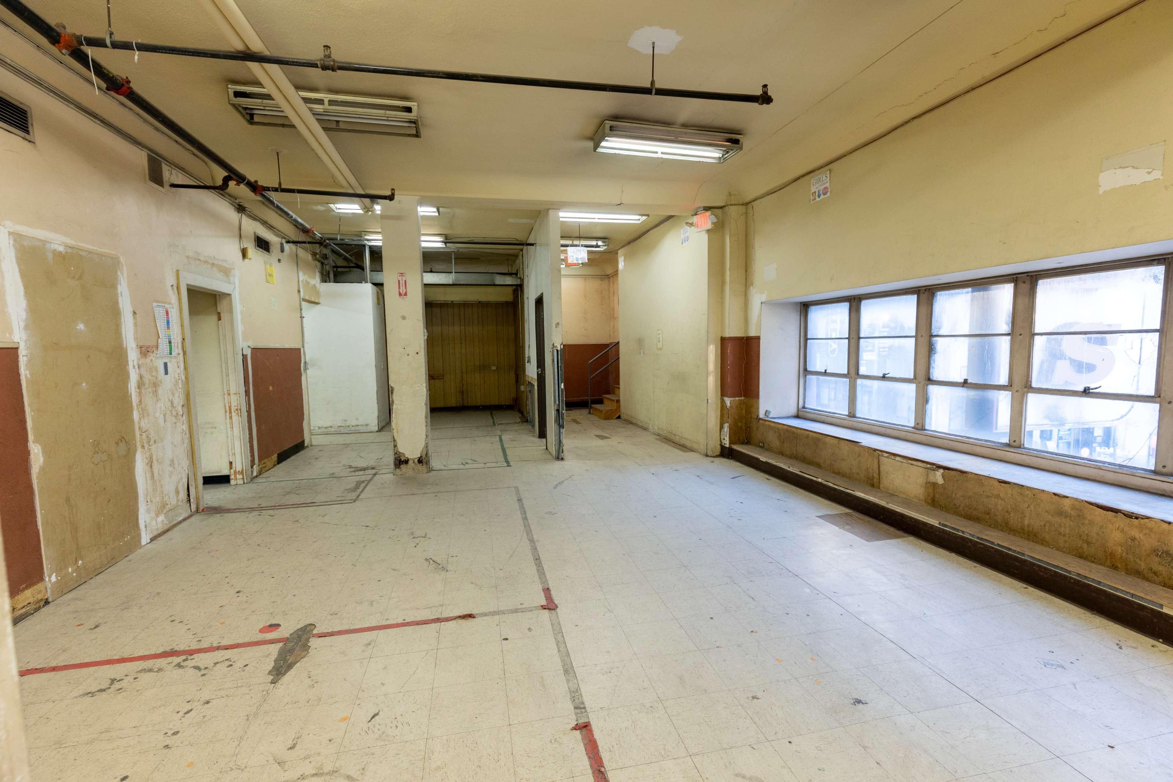 The image shows an empty, partially unfinished interior space with concrete floors, exposed walls, and large windows.