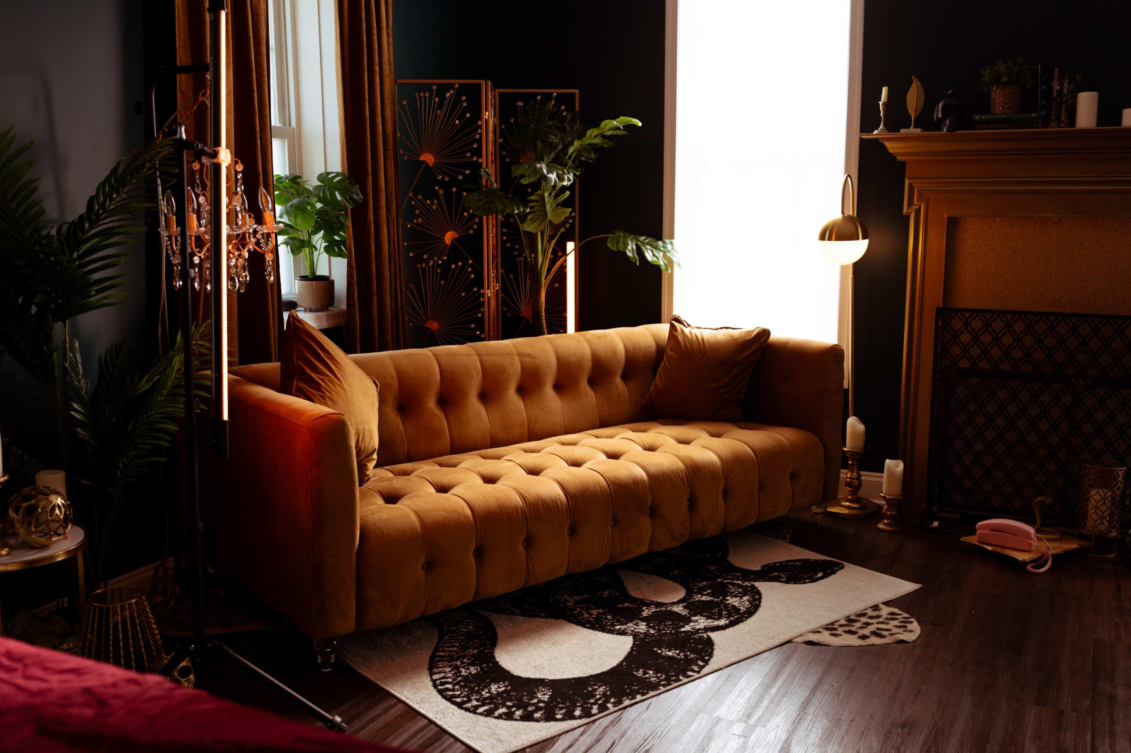 The scene features a stylish living room with a tufted mustard-colored sofa, a decorative screen, various plants, and a cozy fireplace.