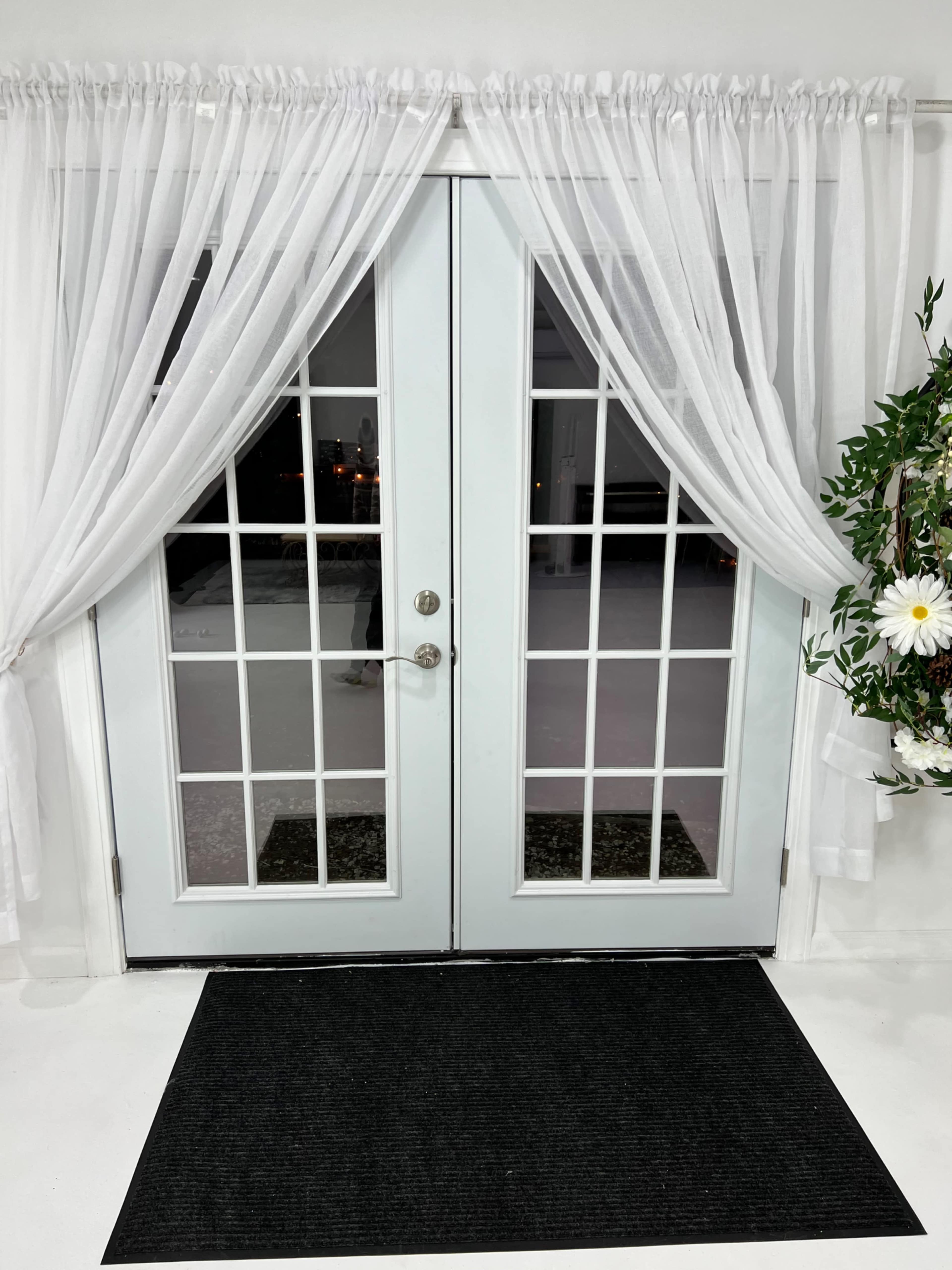 A set of double glass doors adorned with white sheer curtains, flanked by greenery and a welcome mat at the entrance.
