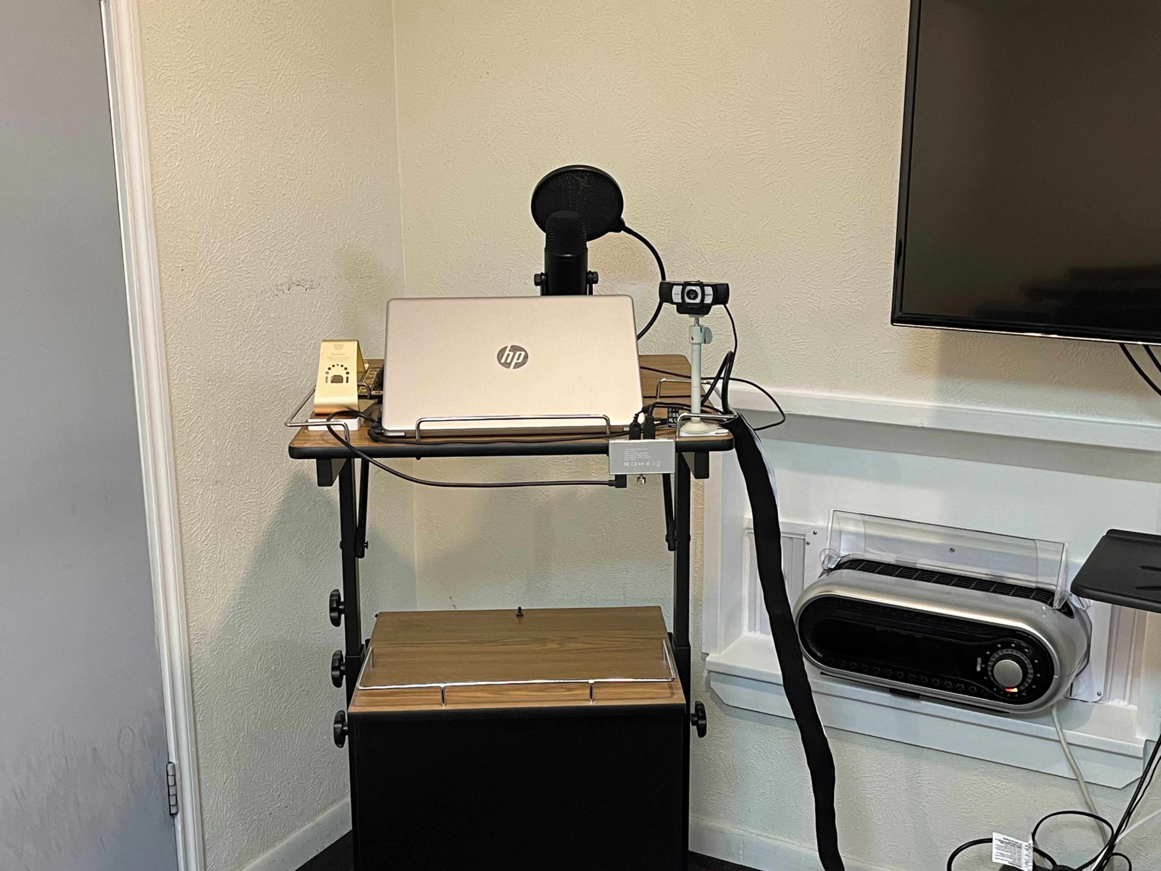 A workspace with a laptop on a wooden cart, a microphone attached, and an electric heater mounted on the wall nearby.