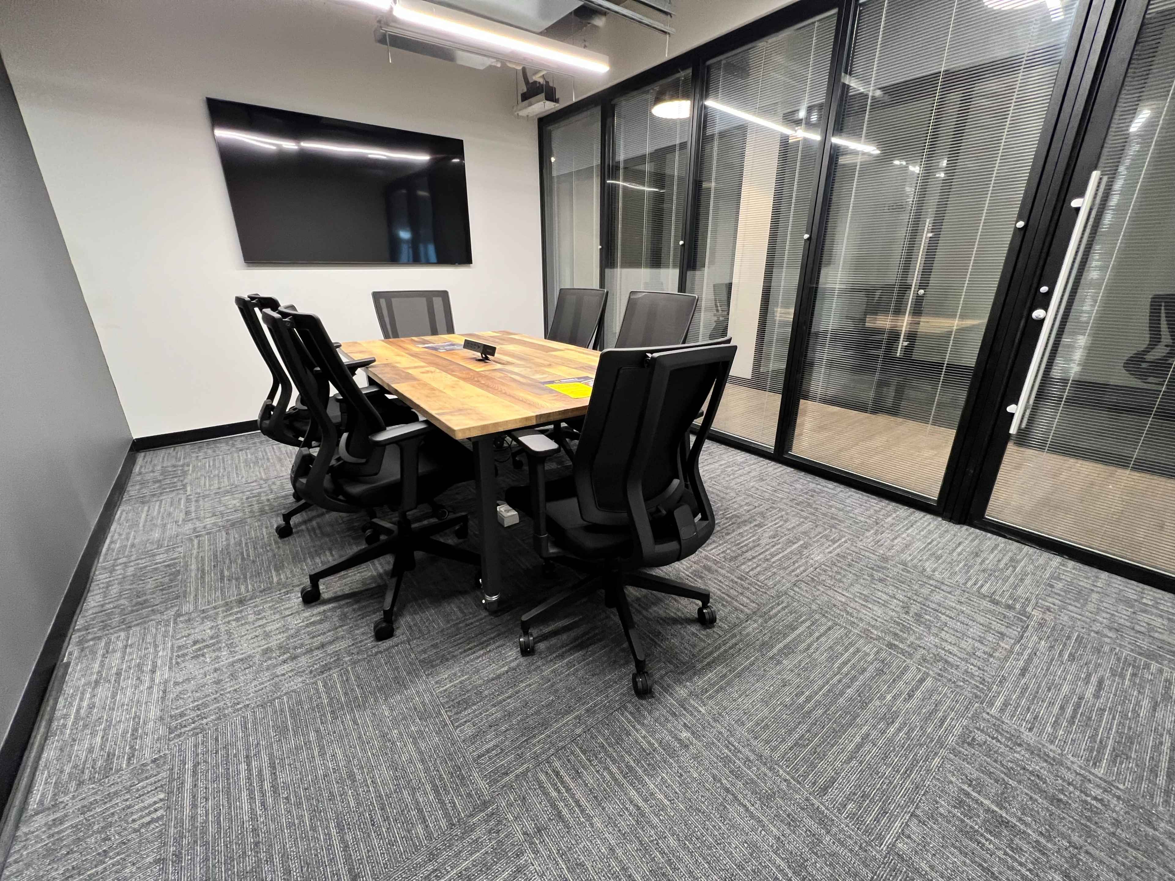 The image shows a modern conference room with a rectangular wooden table and six black ergonomic chairs, surrounded by glass walls and equipped with a wall-mounted screen.