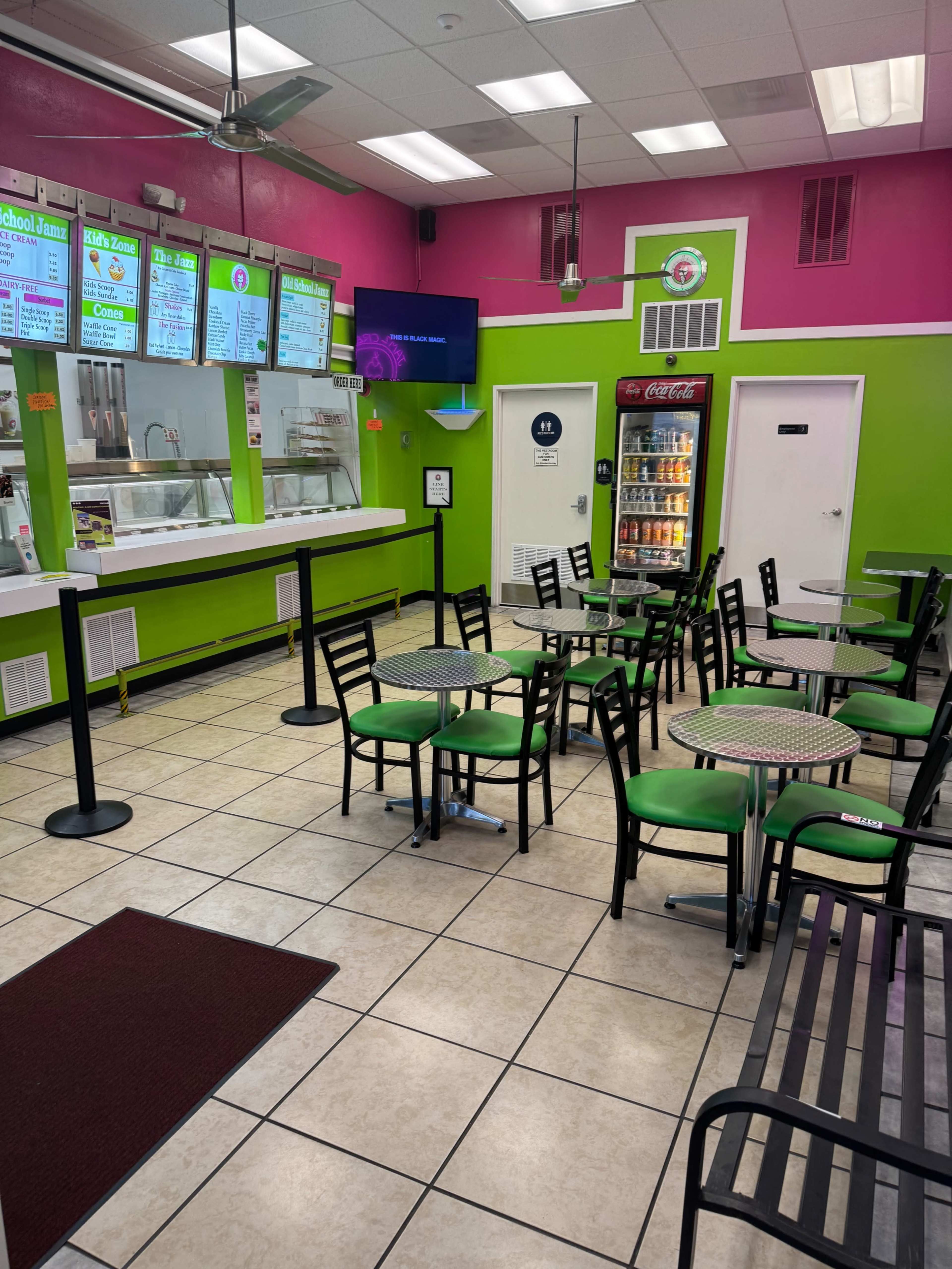 The image shows a brightly colored fast-casual restaurant with green and pink walls, featuring several tables and chairs arranged in the dining area.