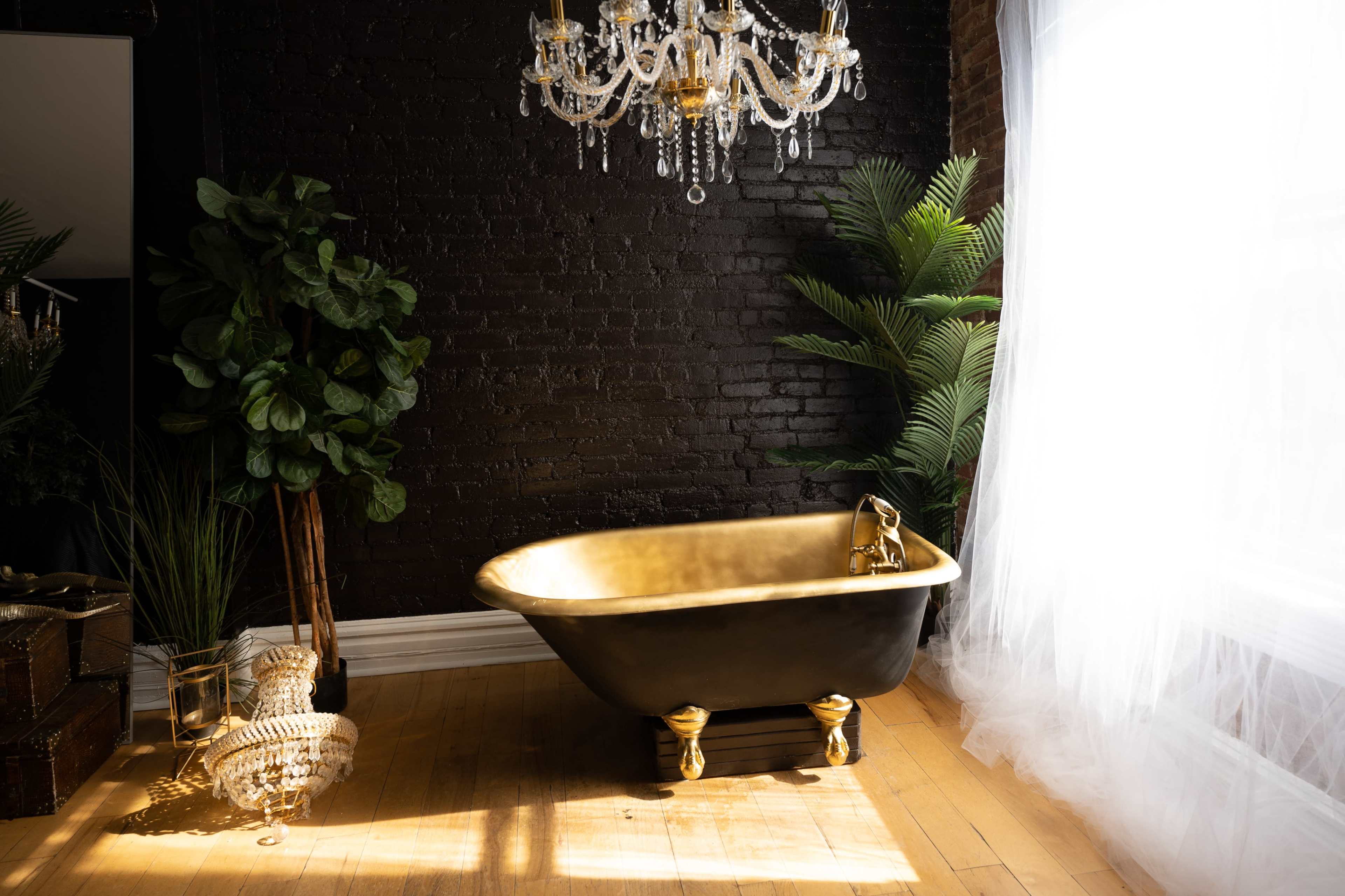 A black and gold freestanding bathtub is situated on hardwood flooring near a large window, surrounded by plants and a crystal chandelier.