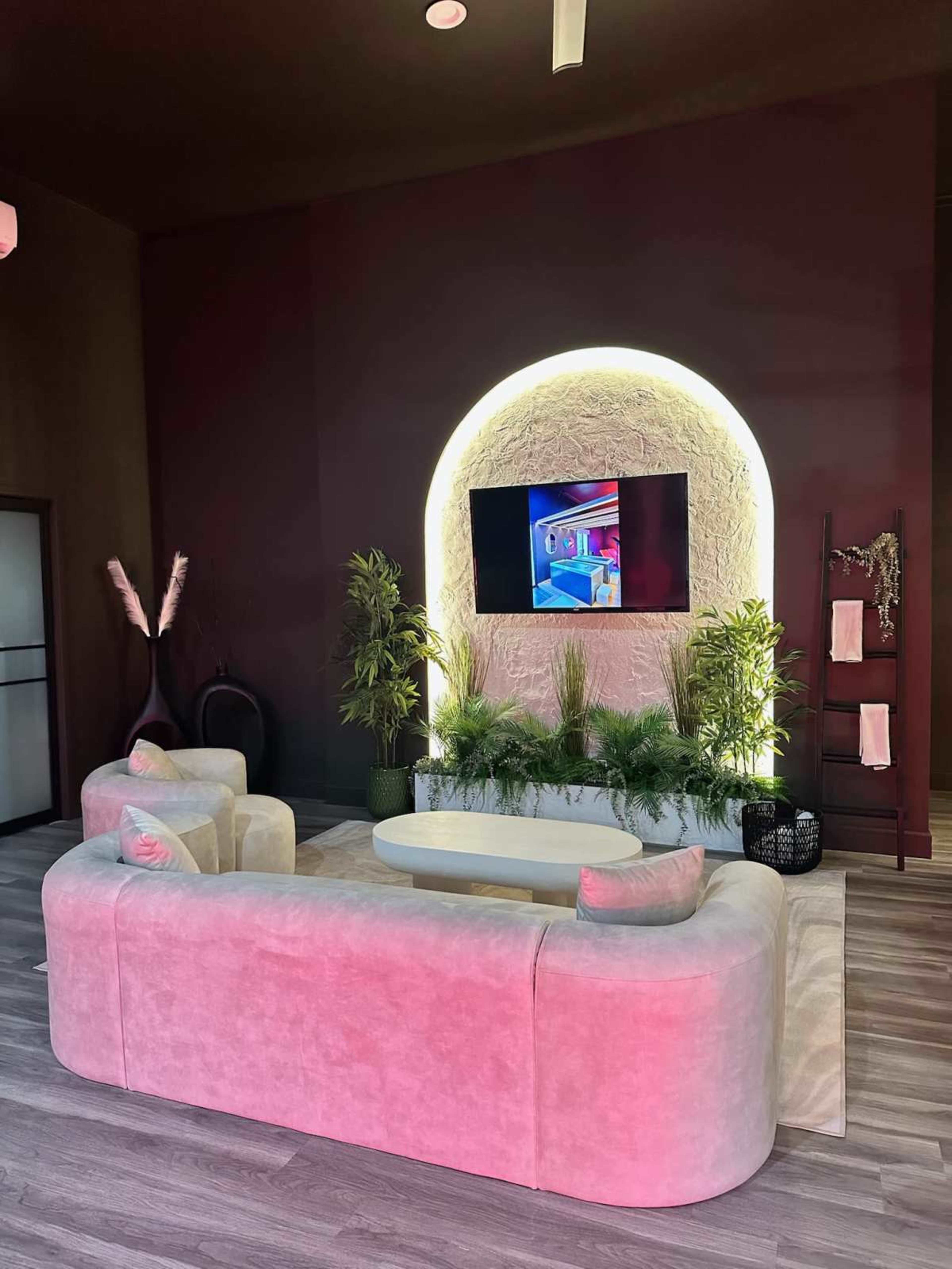 The image shows a modern living room with a curved, light-colored sofa, a circular TV mounted on a textured wall, and decorative plants surrounding the setup.