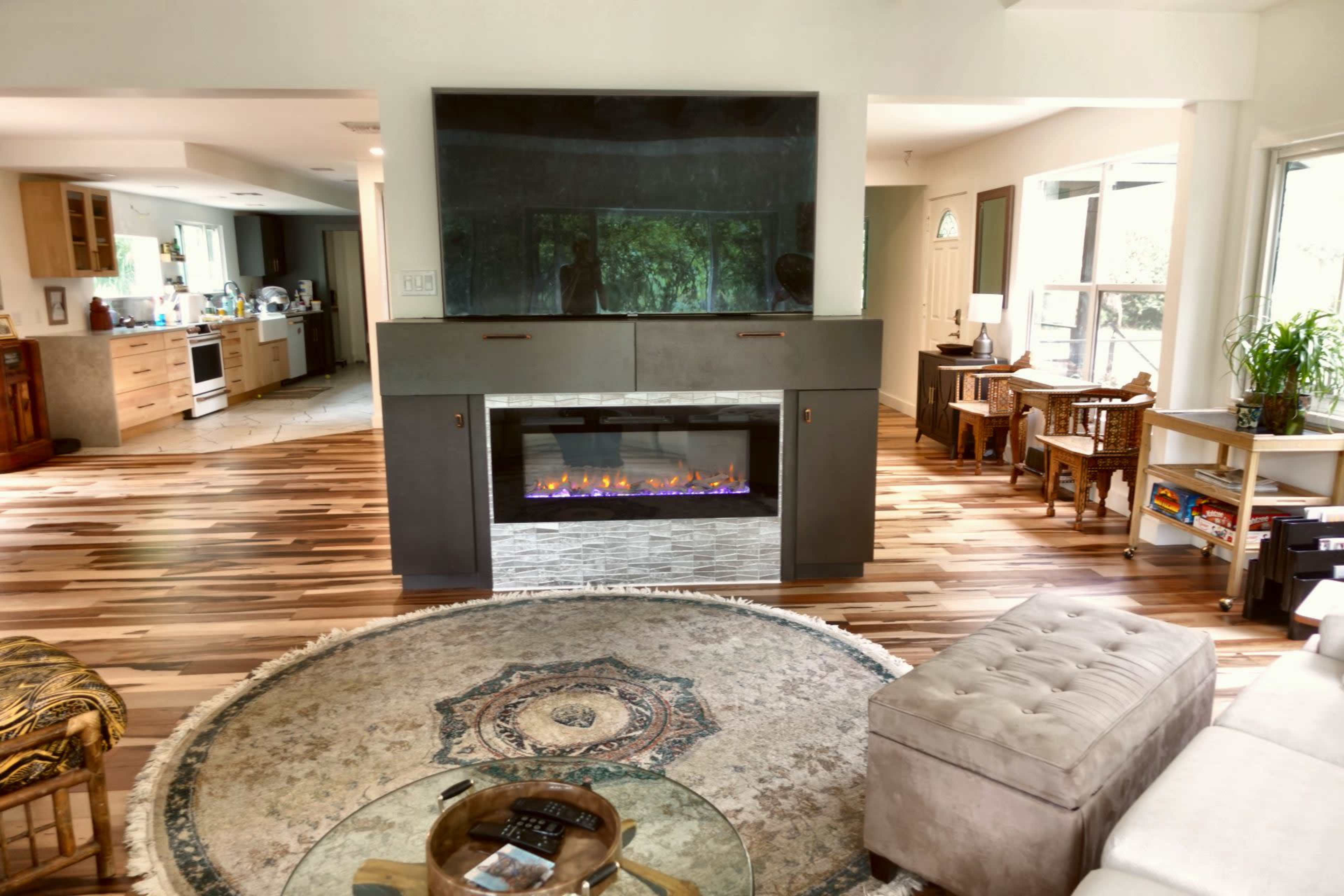 The image shows a spacious living area featuring a modern fireplace, a circular rug, and a view into a kitchen with wooden flooring and furniture.