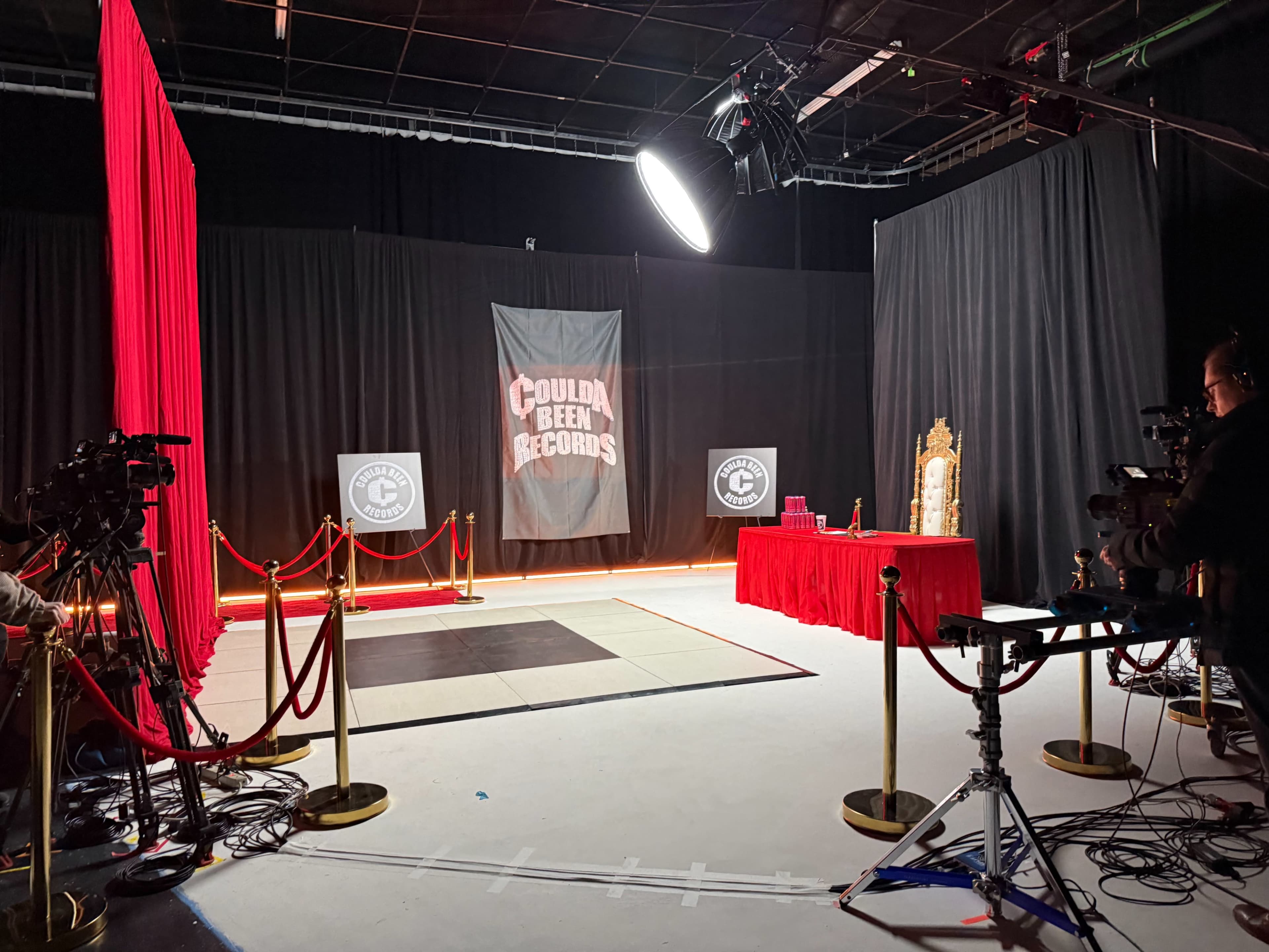 The image shows a film set with a red carpet area, black curtains, camera equipment, and a sign that reads "Coulda Been Records."