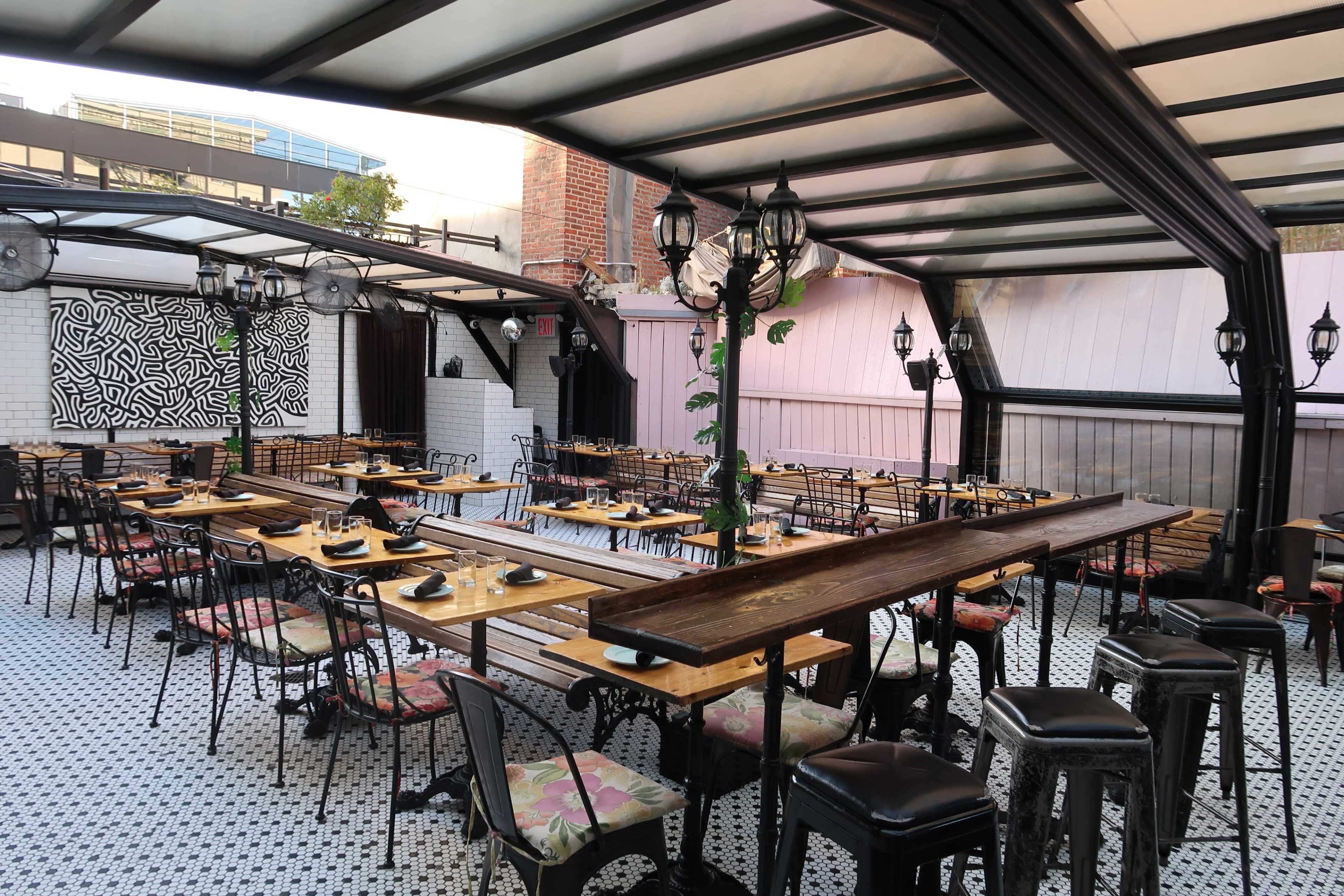 A spacious restaurant patio with wooden tables and metal chairs arranged under a transparent roof, featuring decorative lanterns and a patterned wall in the background.