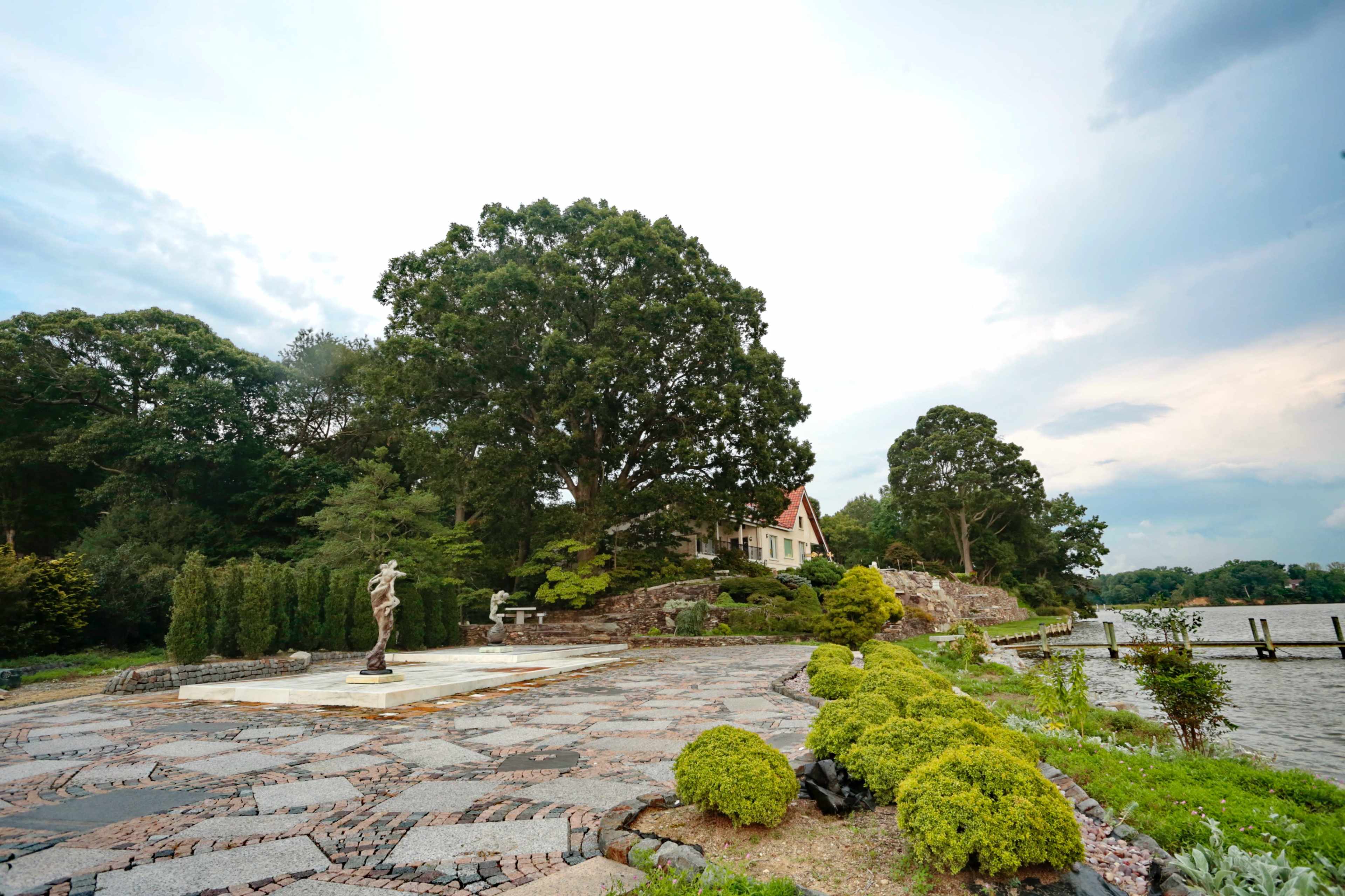 A landscaped waterfront area featuring a stone path, trimmed bushes, a statue, and a house set against a tree-lined background.