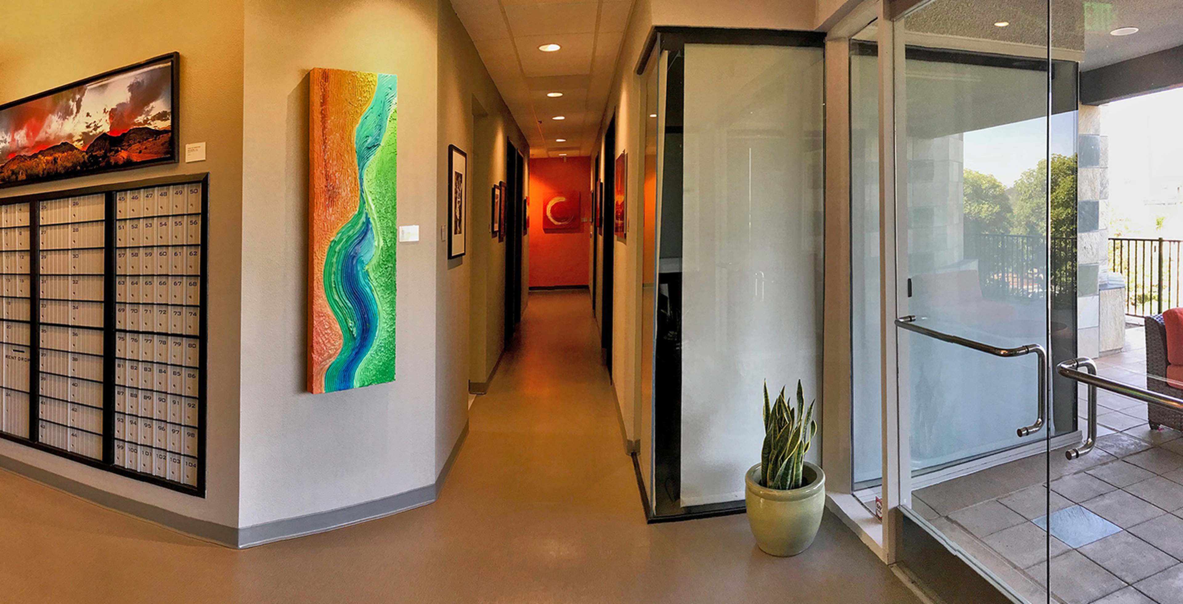 A corridor with mailboxes on one side and colorful artwork on the wall, leading to a glass door that opens to a patio area.