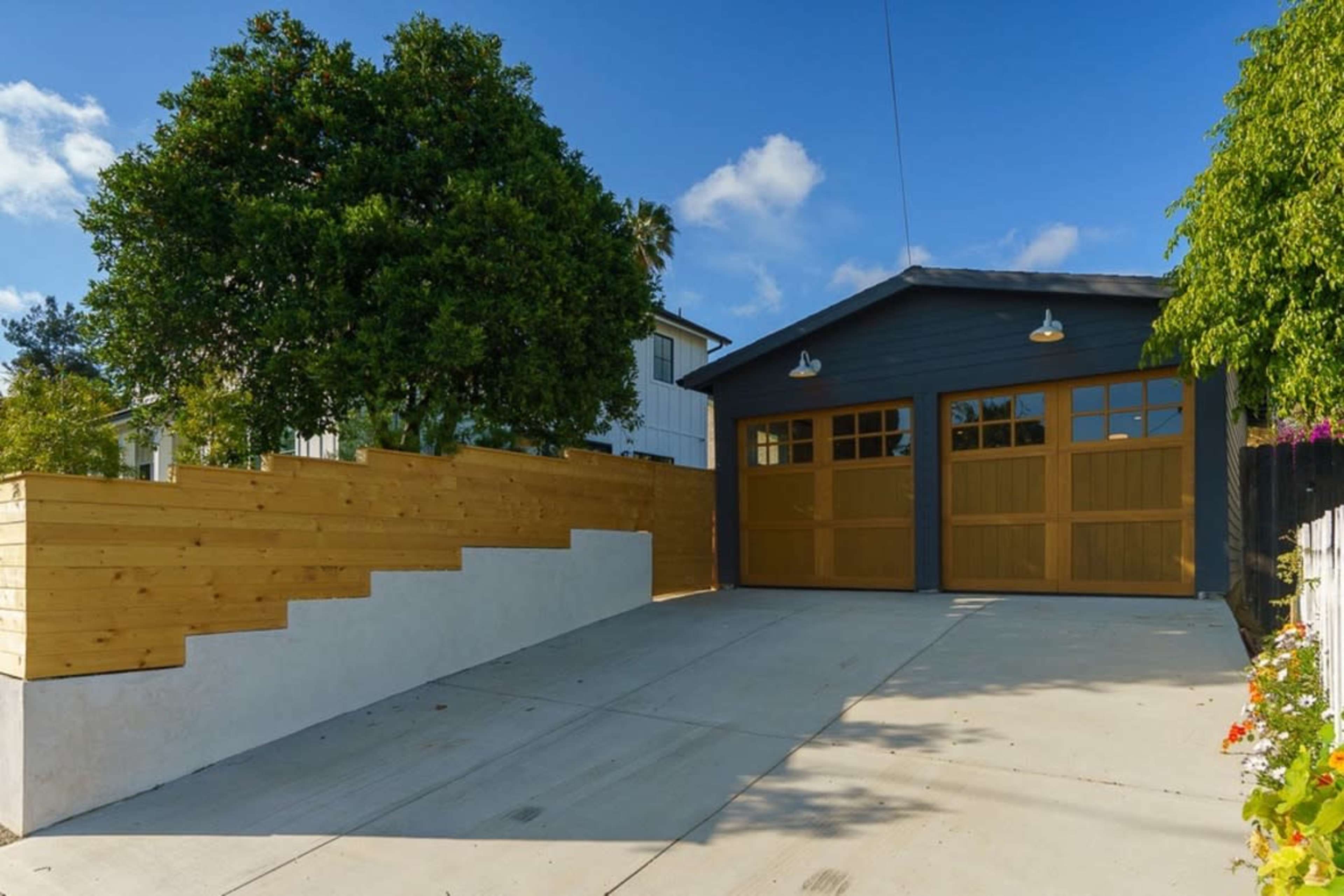 A modern garage with wooden doors is set beside a landscaped driveway featuring a sloped wooden wall and green foliage.
