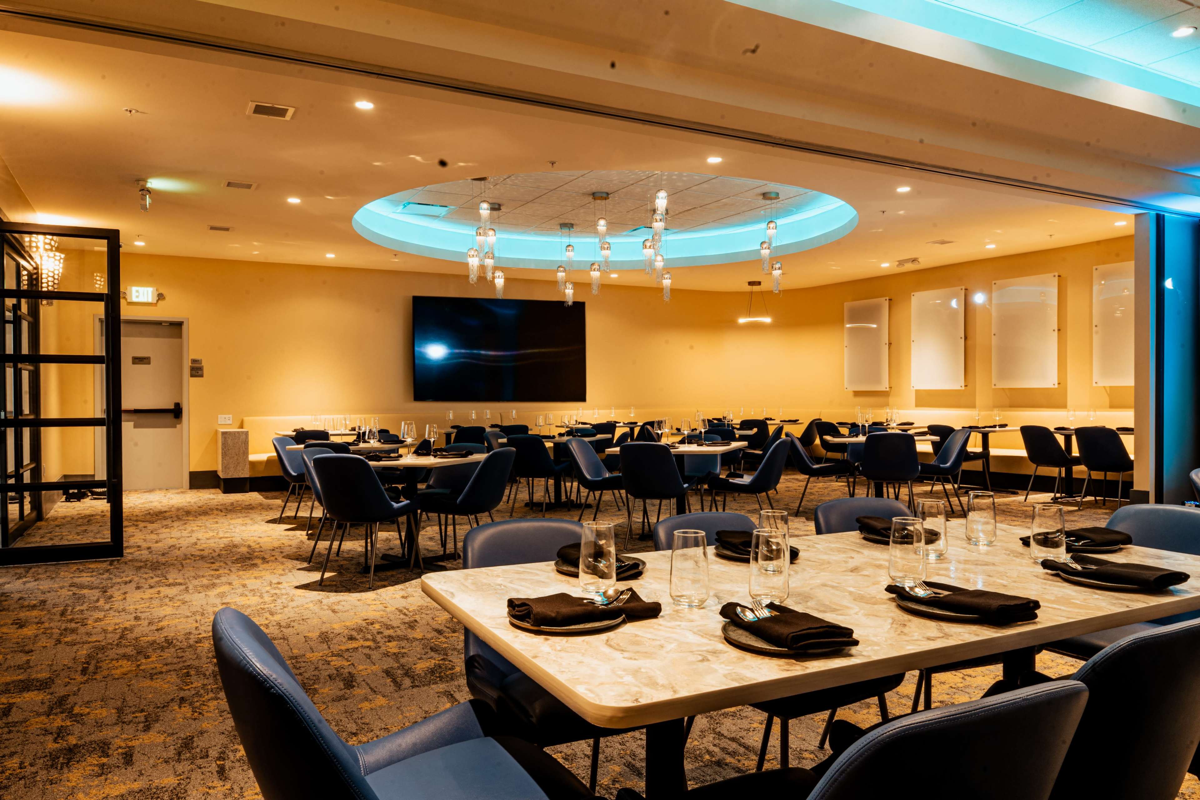 The image shows a modern dining area with blue chairs, round tables, and ambient lighting, featuring a large screen on the wall and decorative ceiling fixtures.