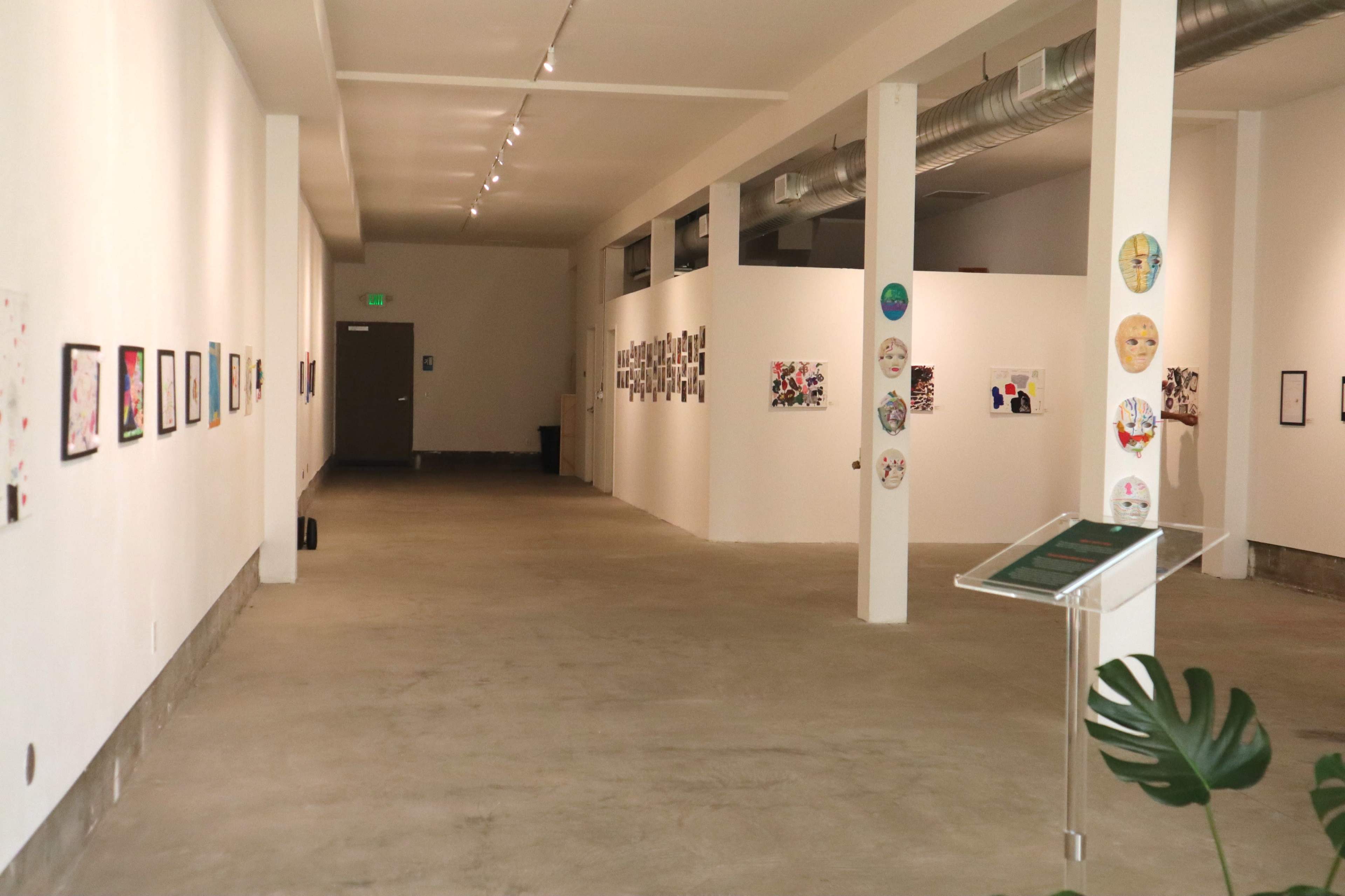 The image shows a gallery interior with white walls, concrete flooring, and artwork displayed along the left side.