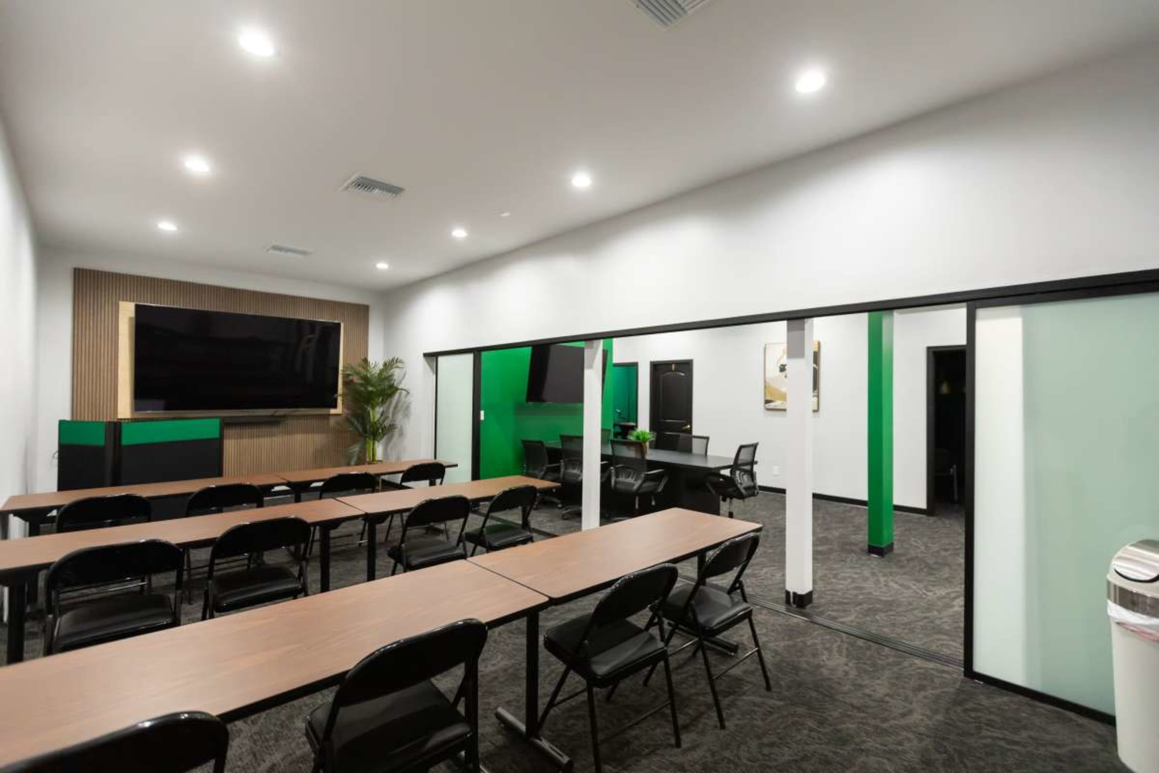 The image shows a modern meeting room with a series of long tables and chairs arranged facing a large screen and wall-mounted television.