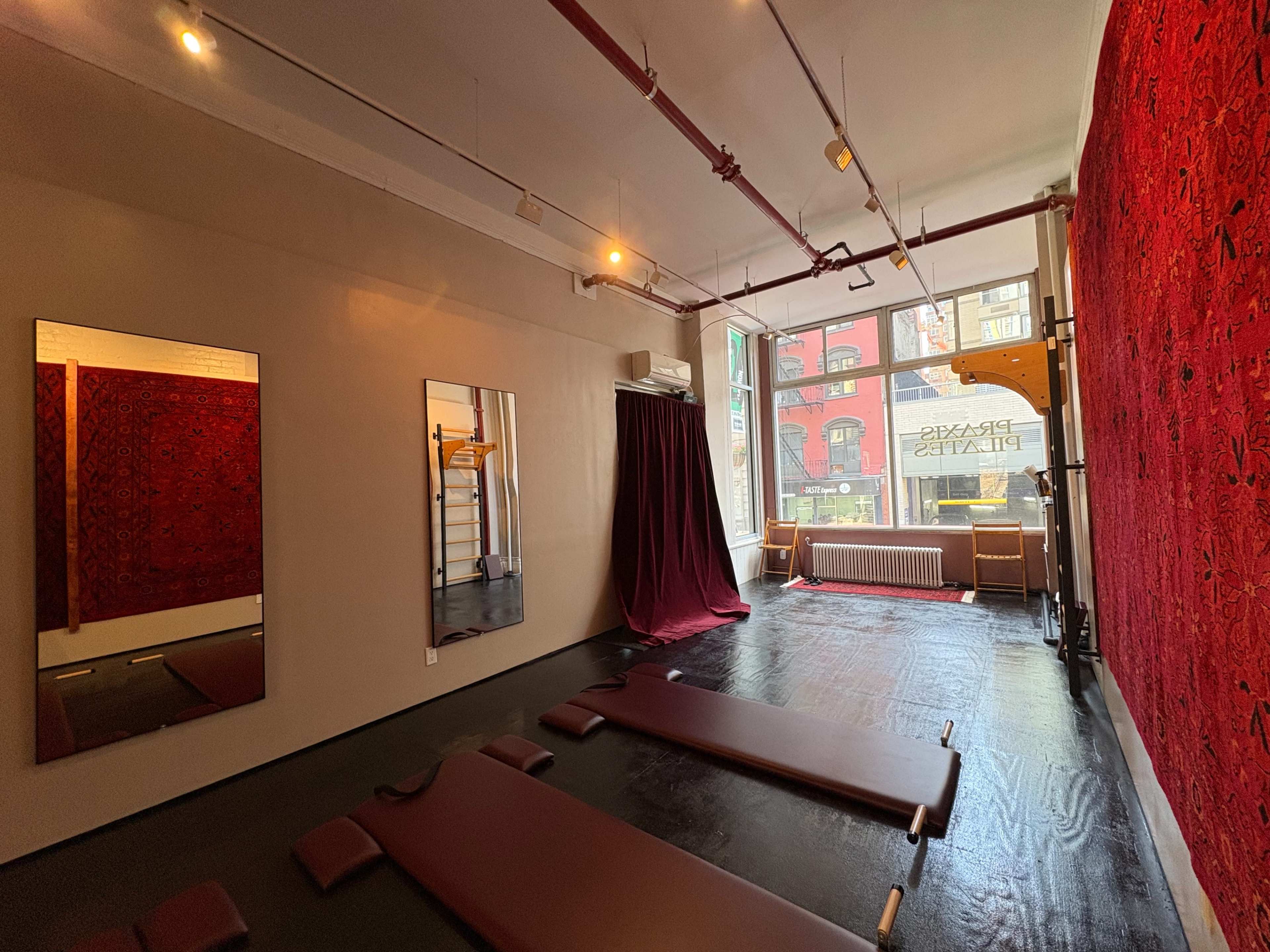 The image shows a spacious, minimalist room designed for yoga or Pilates, featuring wooden floors, mirrors, two exercise mats, and decorative red wall hangings.