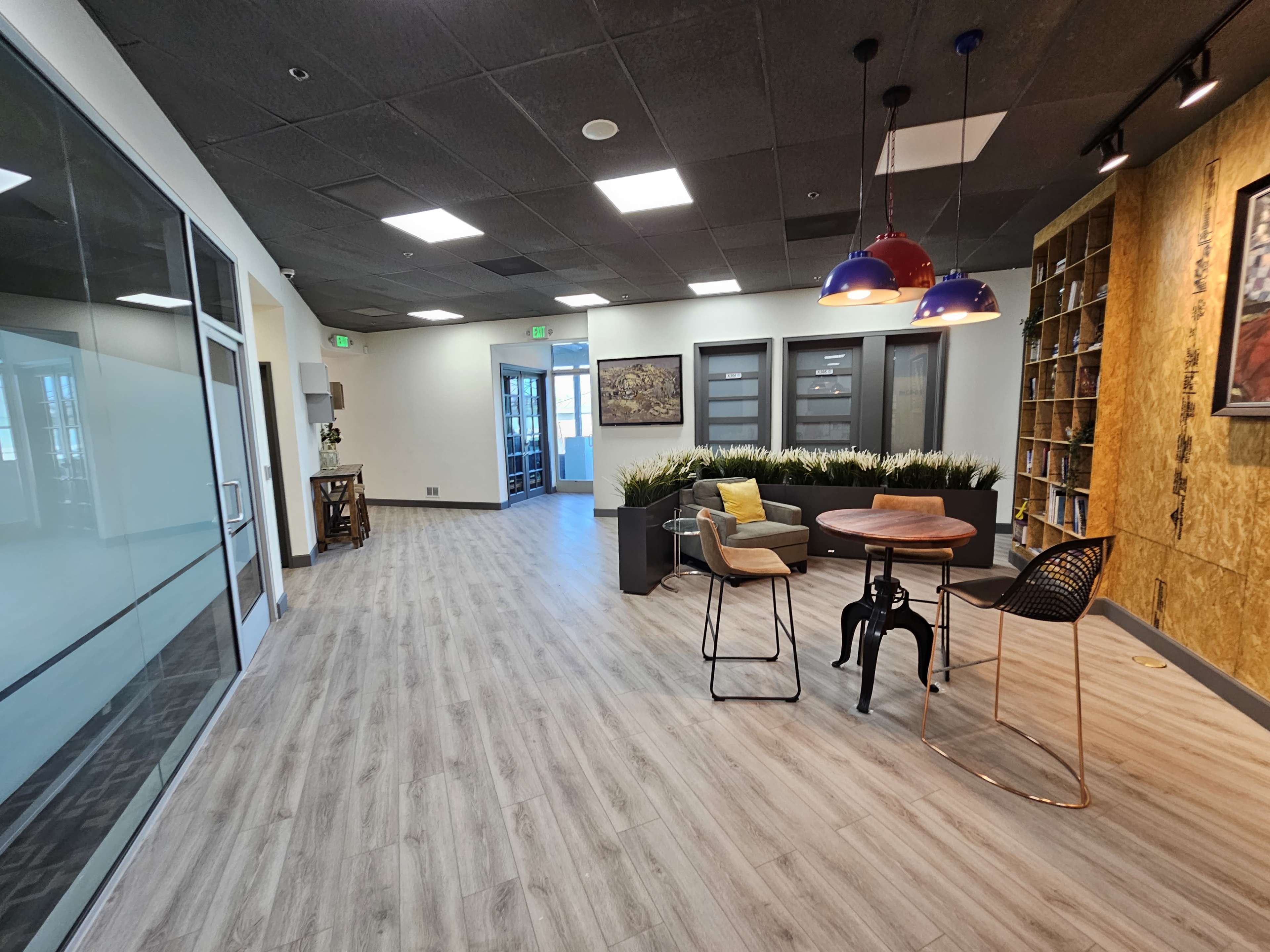 A modern office lobby with sleek flooring, a seating area featuring a round table and chairs, and large windows leading to an adjacent room.