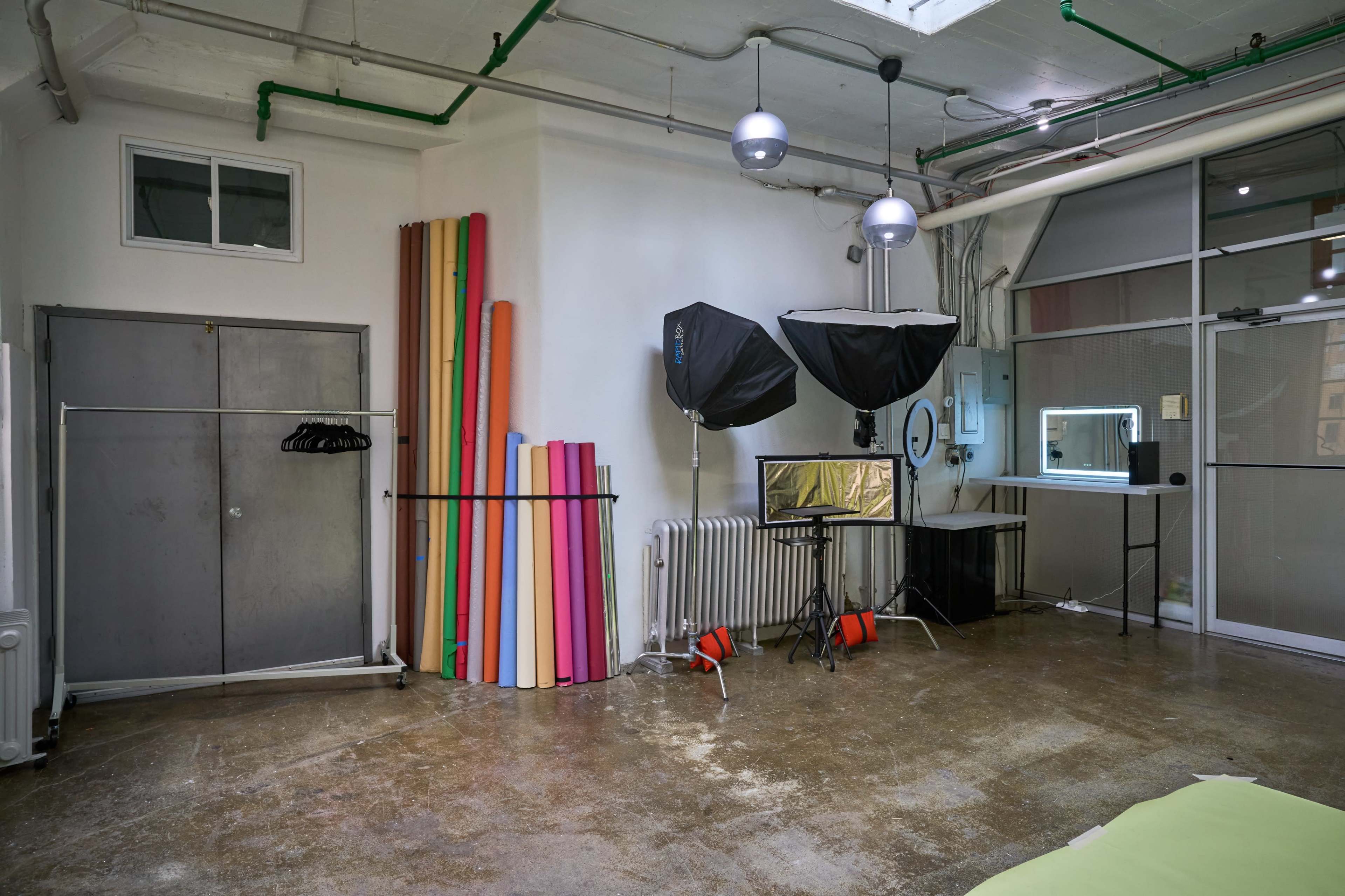 A well-lit studio space featuring colorful backdrop rolls, a photography setup with softboxes, and a large window.