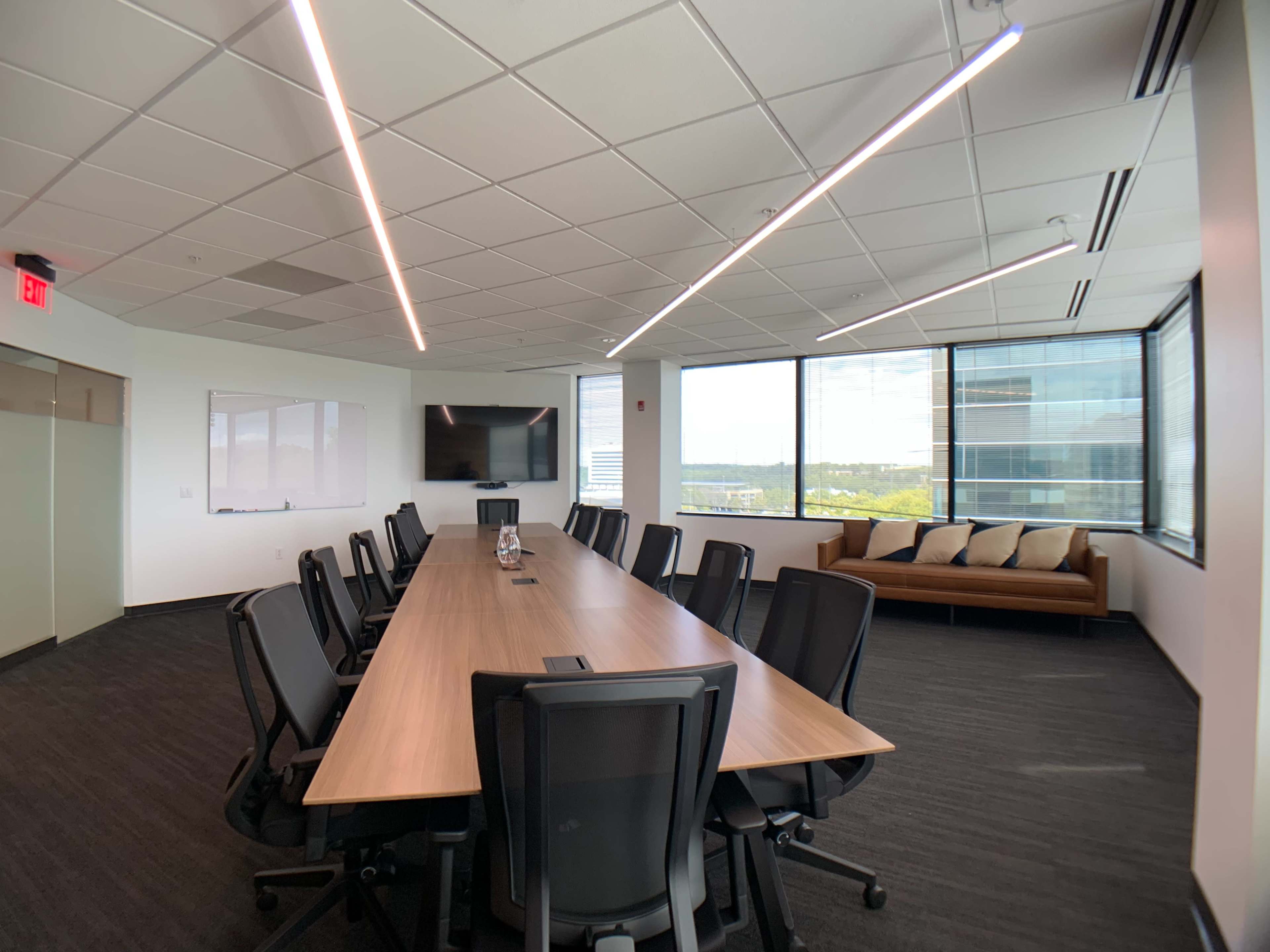 A modern conference room features a long wooden table surrounded by black chairs, with large windows offering a view of the outside.