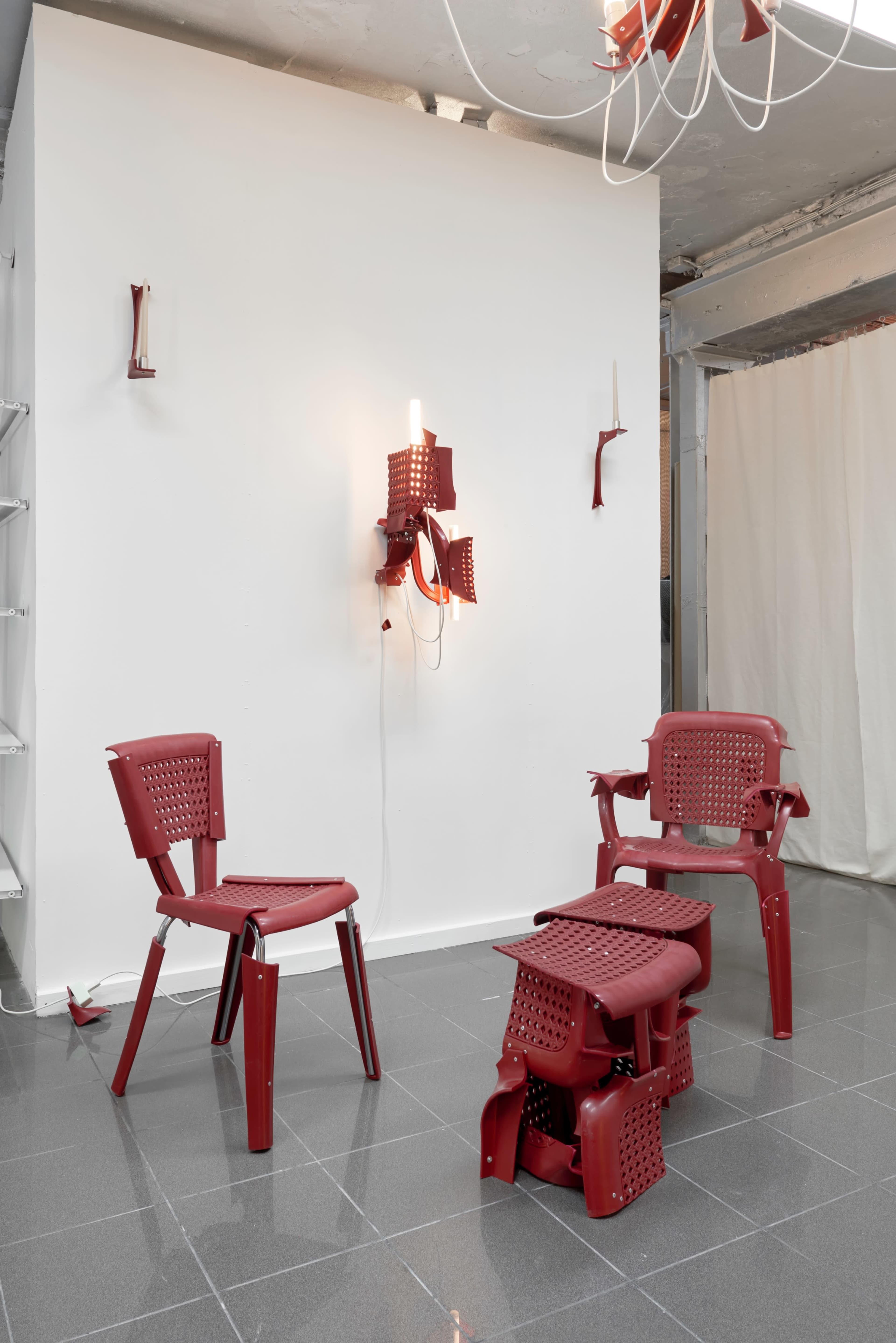 The image shows a minimalist indoor space featuring three red plastic chairs and a red wall-mounted light installation against a white wall.