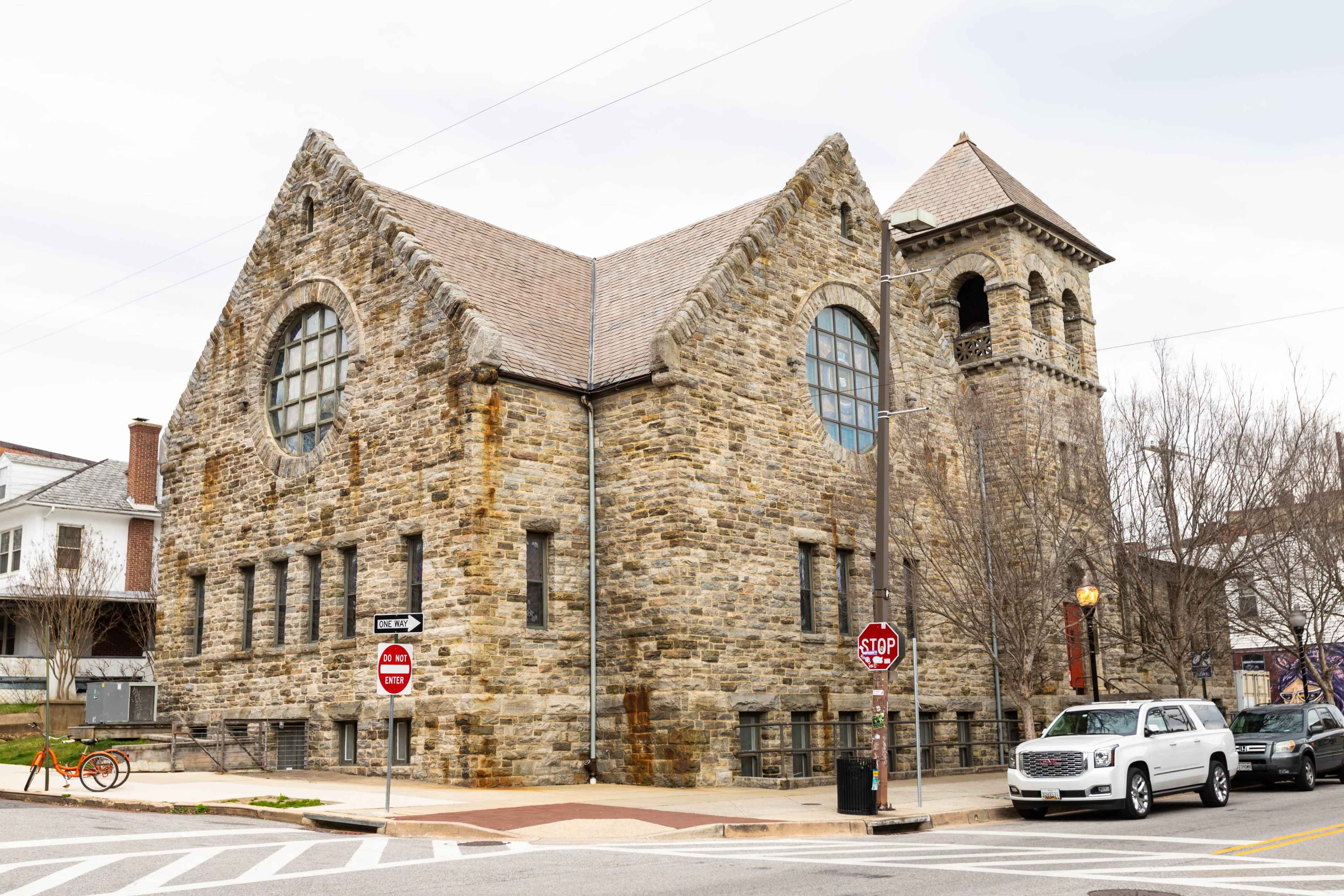 The image shows a large, stone building with a distinctive bell tower and a gabled roof located at a street intersection.
