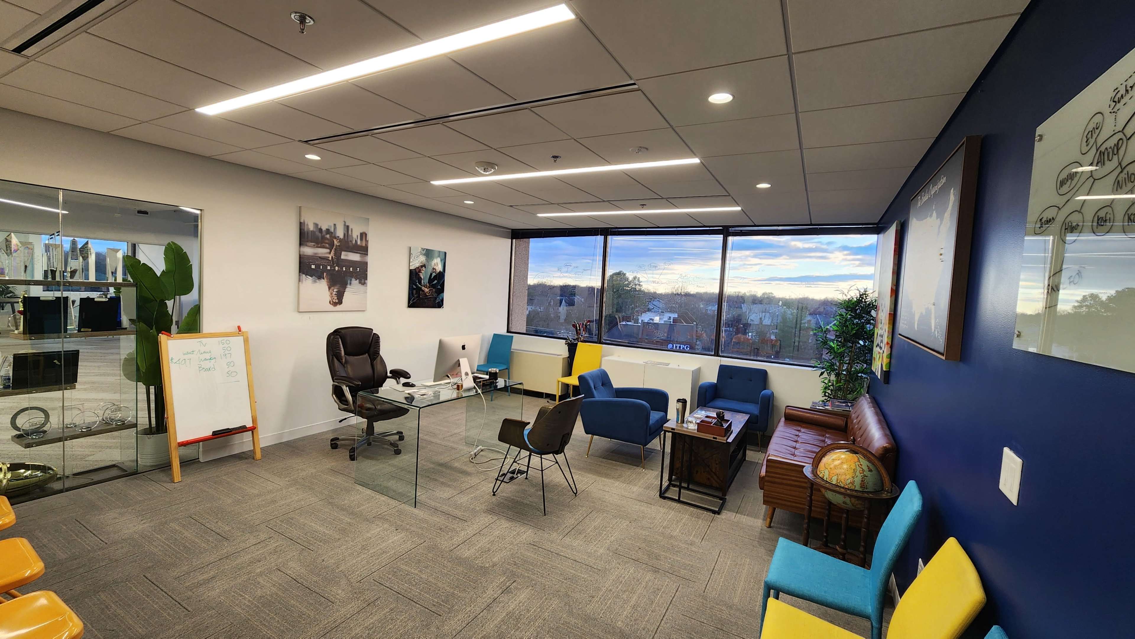 A modern office space featuring glass walls, a desk with a computer, a black office chair, colorful seating, and large windows overlooking a scenic view.