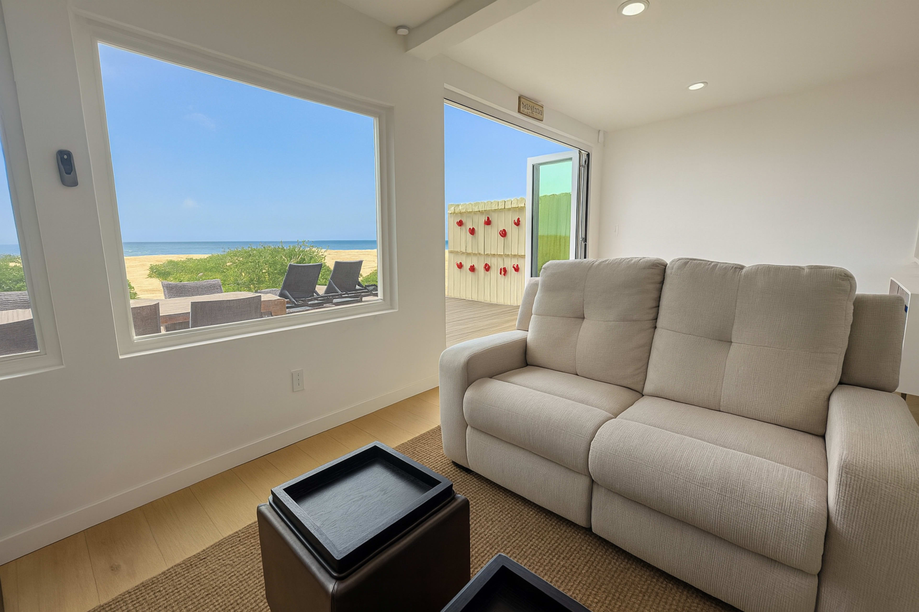 A light-filled room features a beige couch and large windows that open to a view of a sandy beach and the ocean.