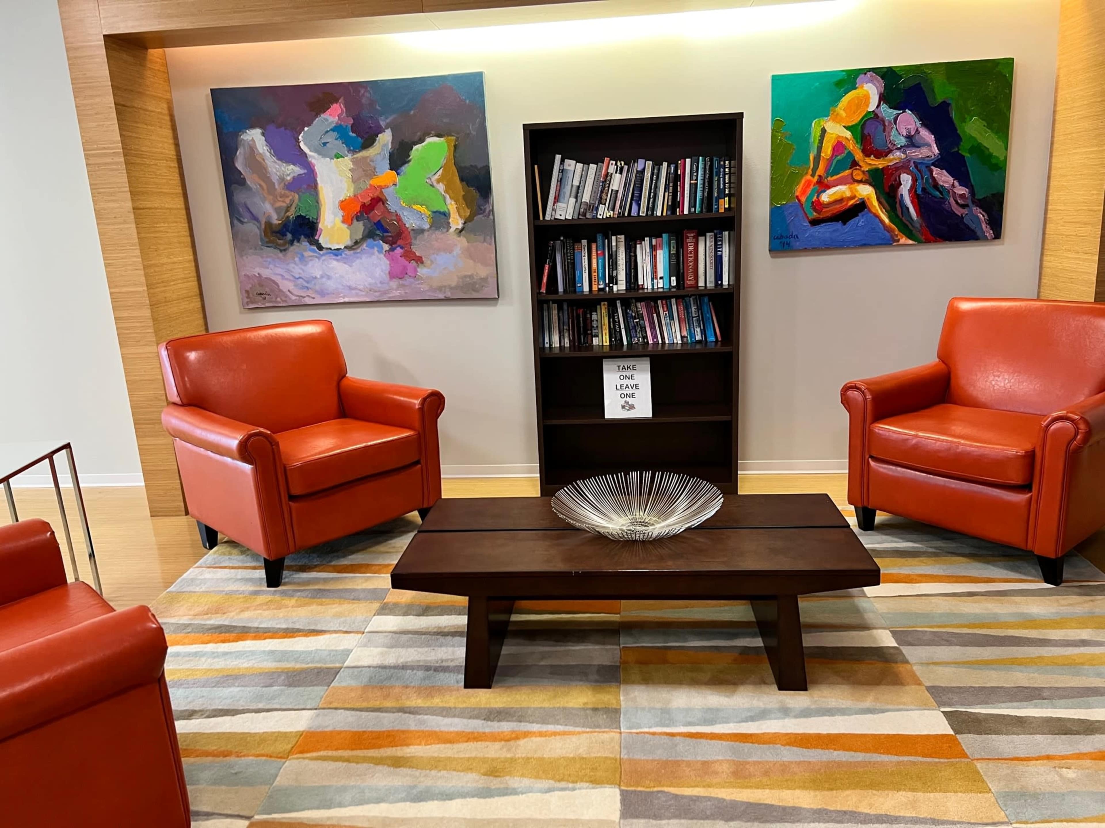 A cozy seating area with two orange armchairs facing a low wooden coffee table, with a bookshelf and colorful abstract paintings on the walls.