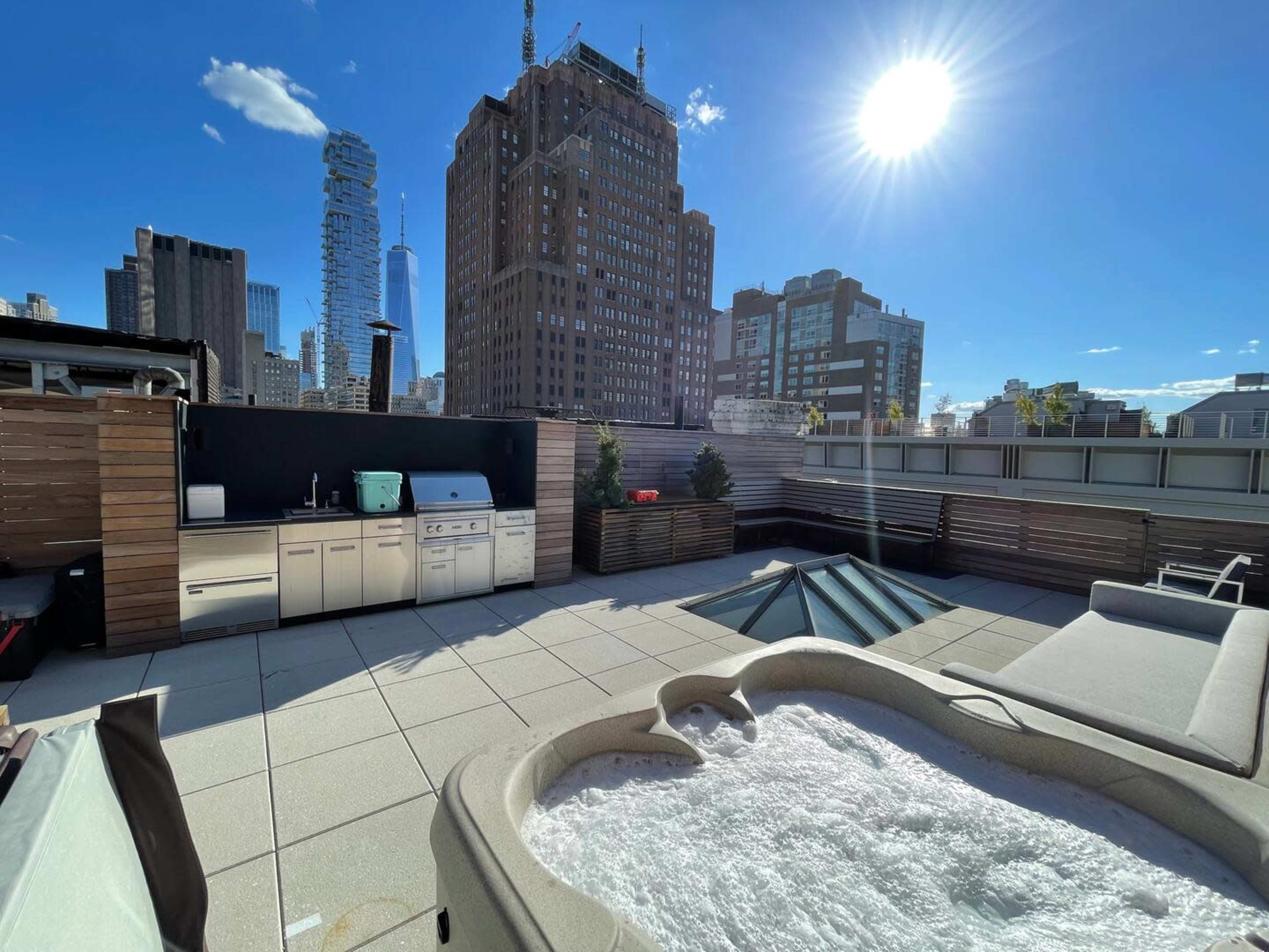 A rooftop space featuring a hot tub, an outdoor kitchen area, and a view of tall city buildings under bright sunlight.