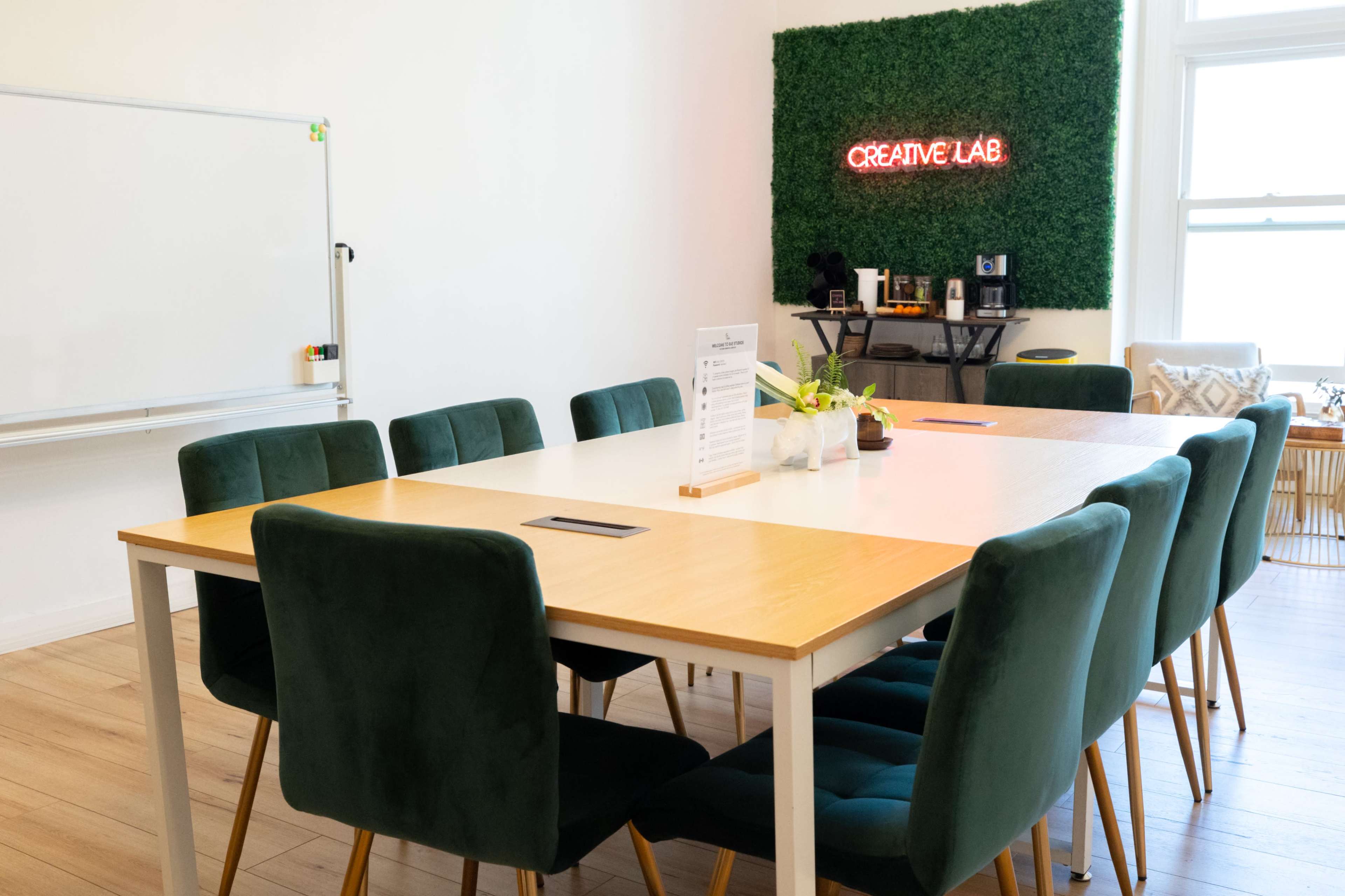 A large wooden table with green chairs is arranged in a bright room featuring a "CREATIVE LAB" sign made of greenery on the wall.