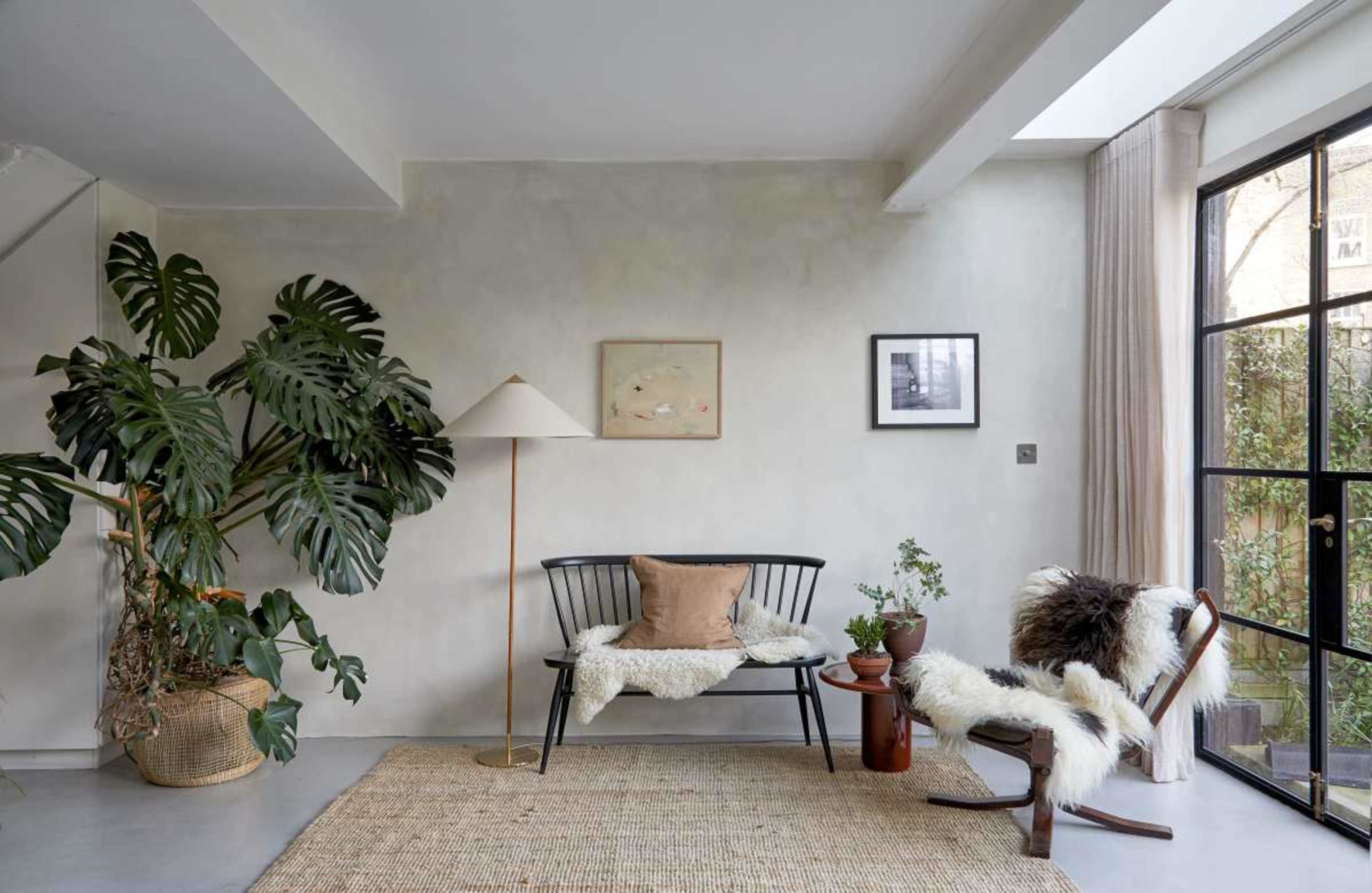 The scene features a cozy, modern living room with a bench, a rocking chair, various plants, and large windows allowing natural light to enter.