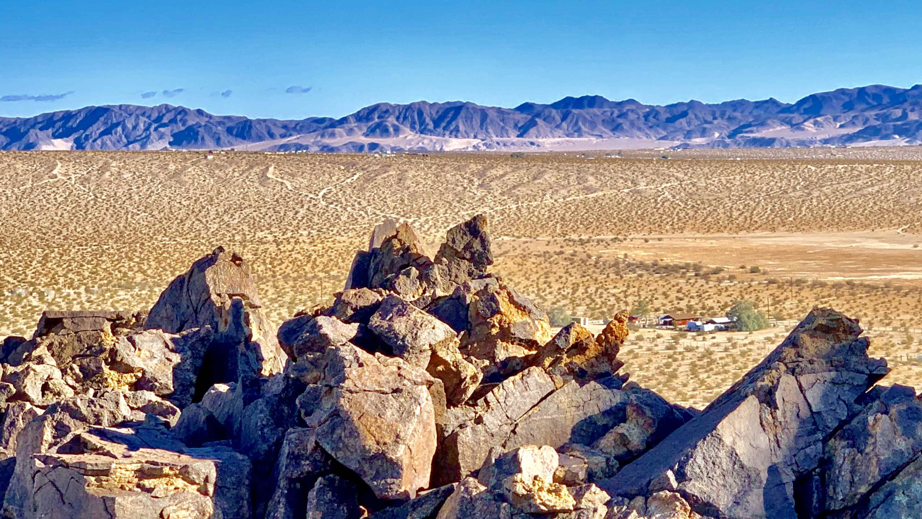A rocky outcrop overlooks a distant settlement surrounded by vast desert and mountains under a clear blue sky.