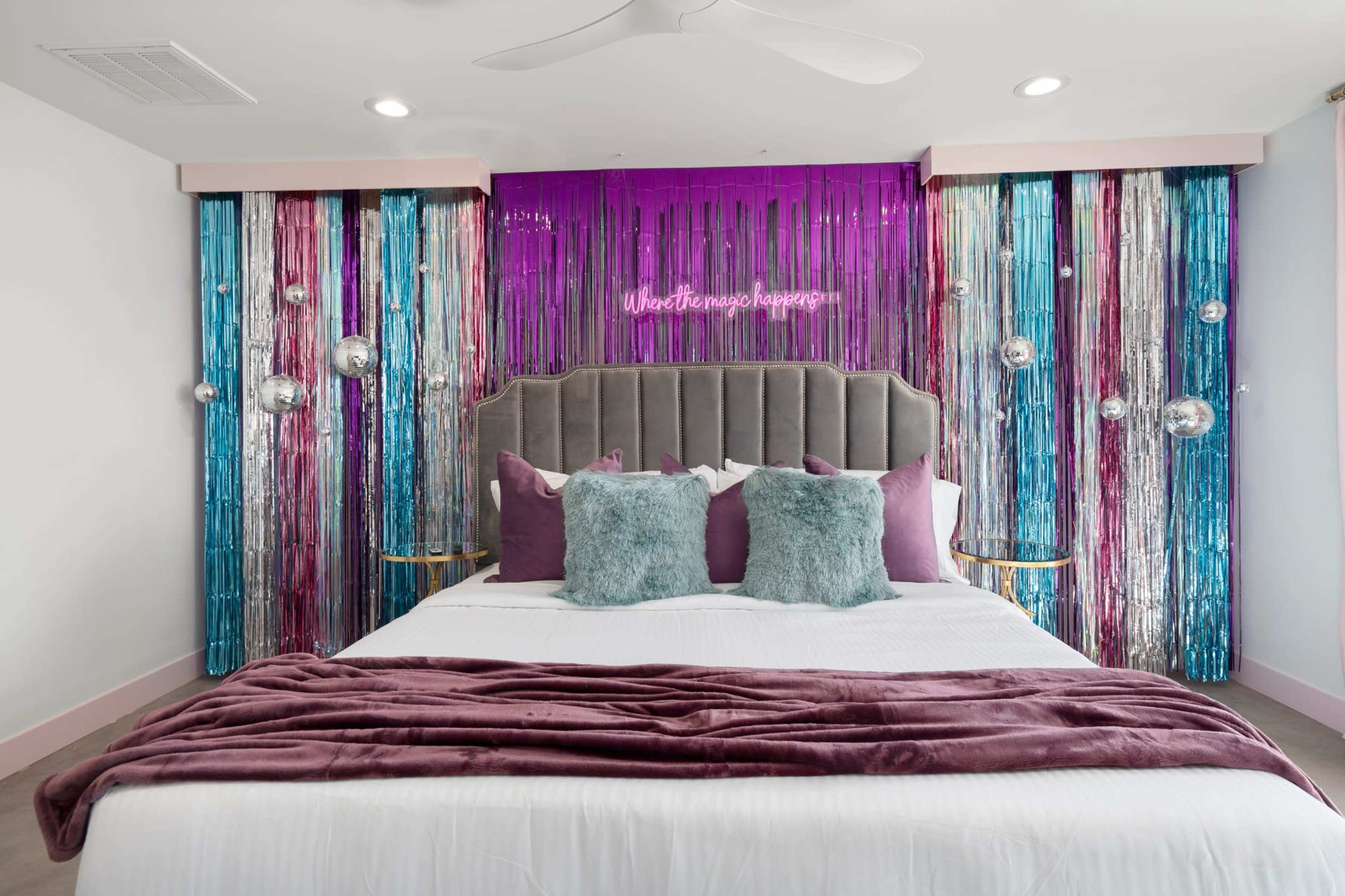 The image shows a modern bedroom with a bed featuring a gray headboard, adorned with purple and teal decorative elements, and a backdrop of shiny streamers and a neon sign.