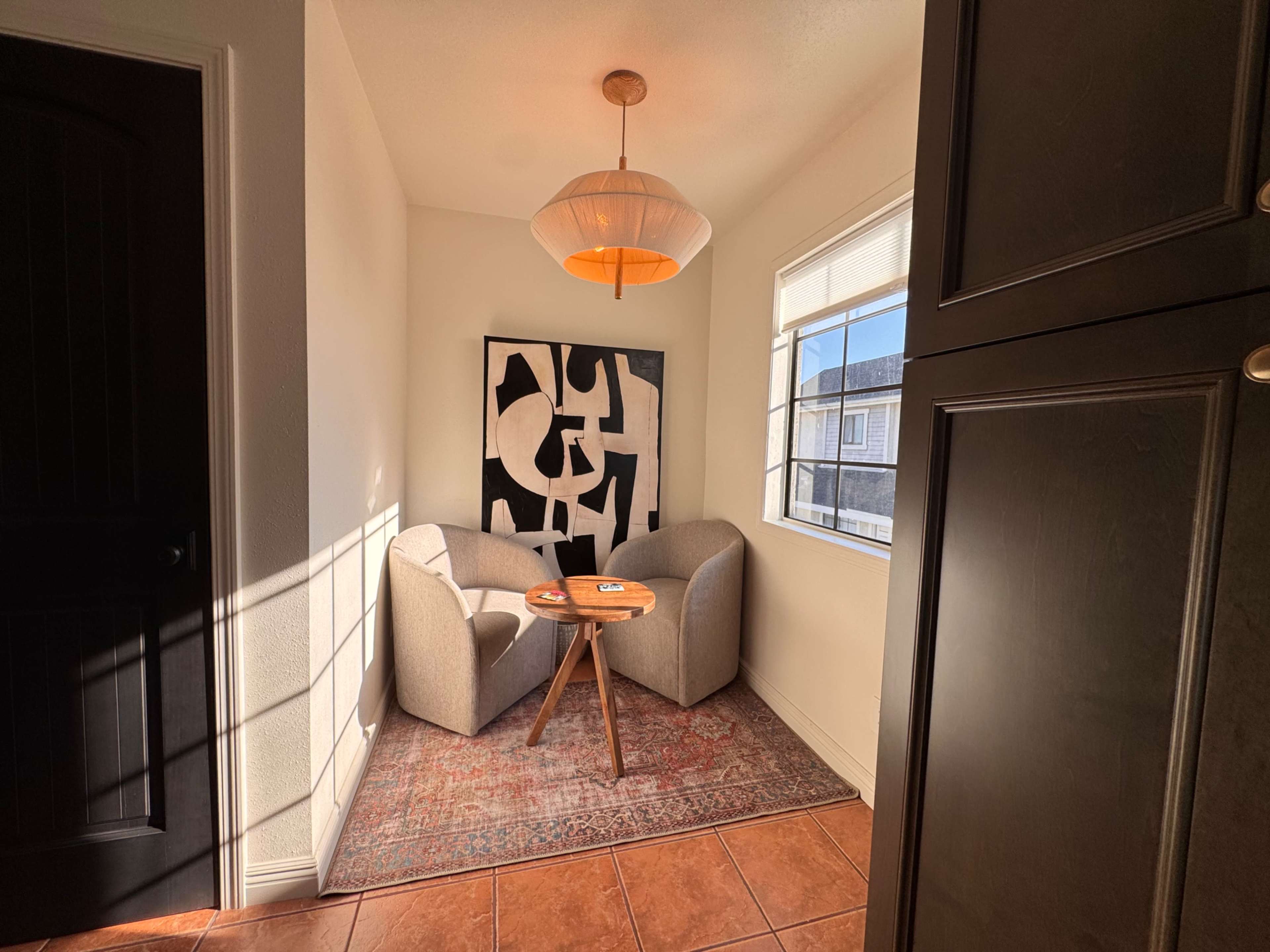 A small, cozy nook features two gray armchairs flanking a wooden side table, with a modern art piece on the wall and natural light streaming in from a nearby window.