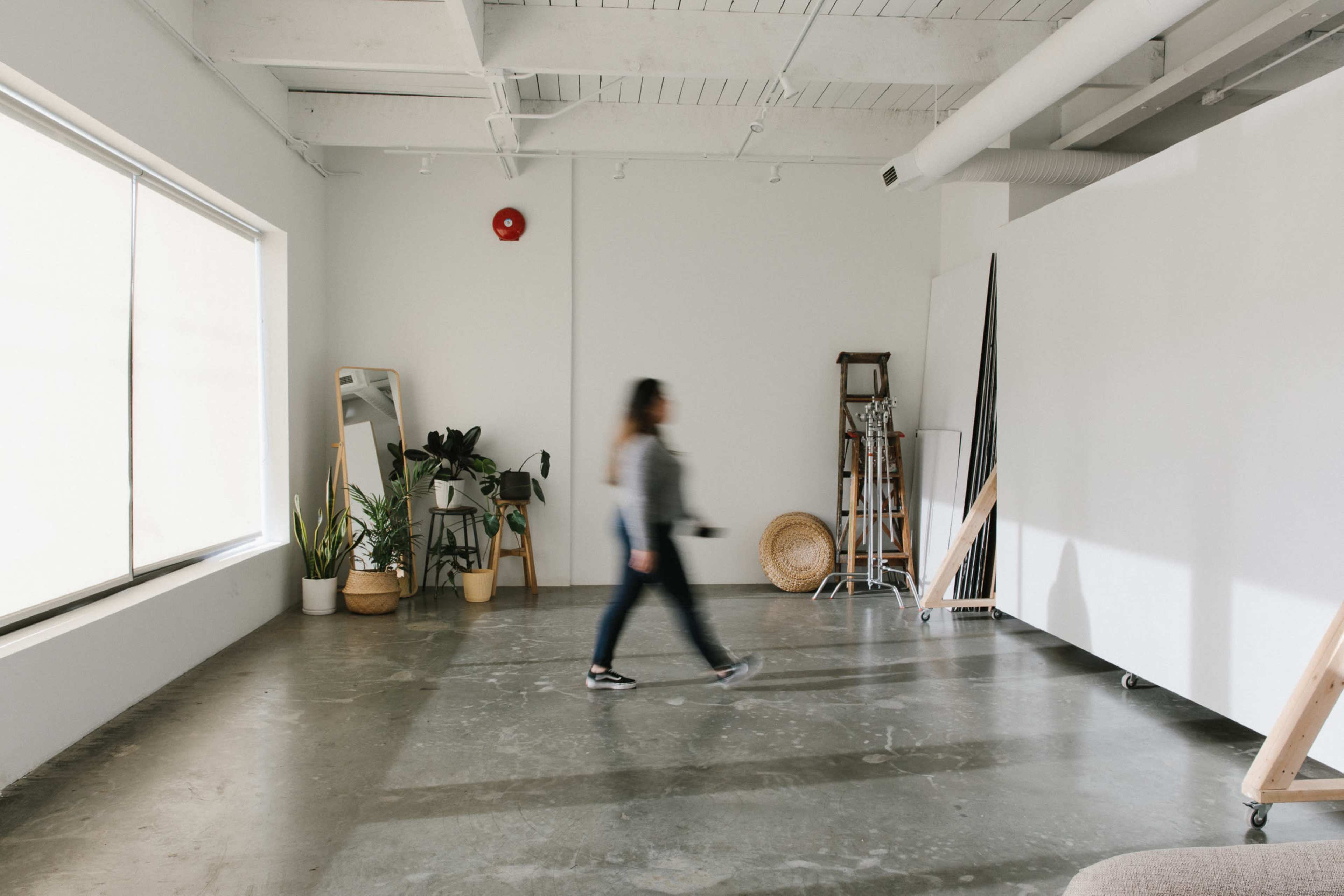 A person walks through a bright, minimalist studio with plants and mirrors along the walls.