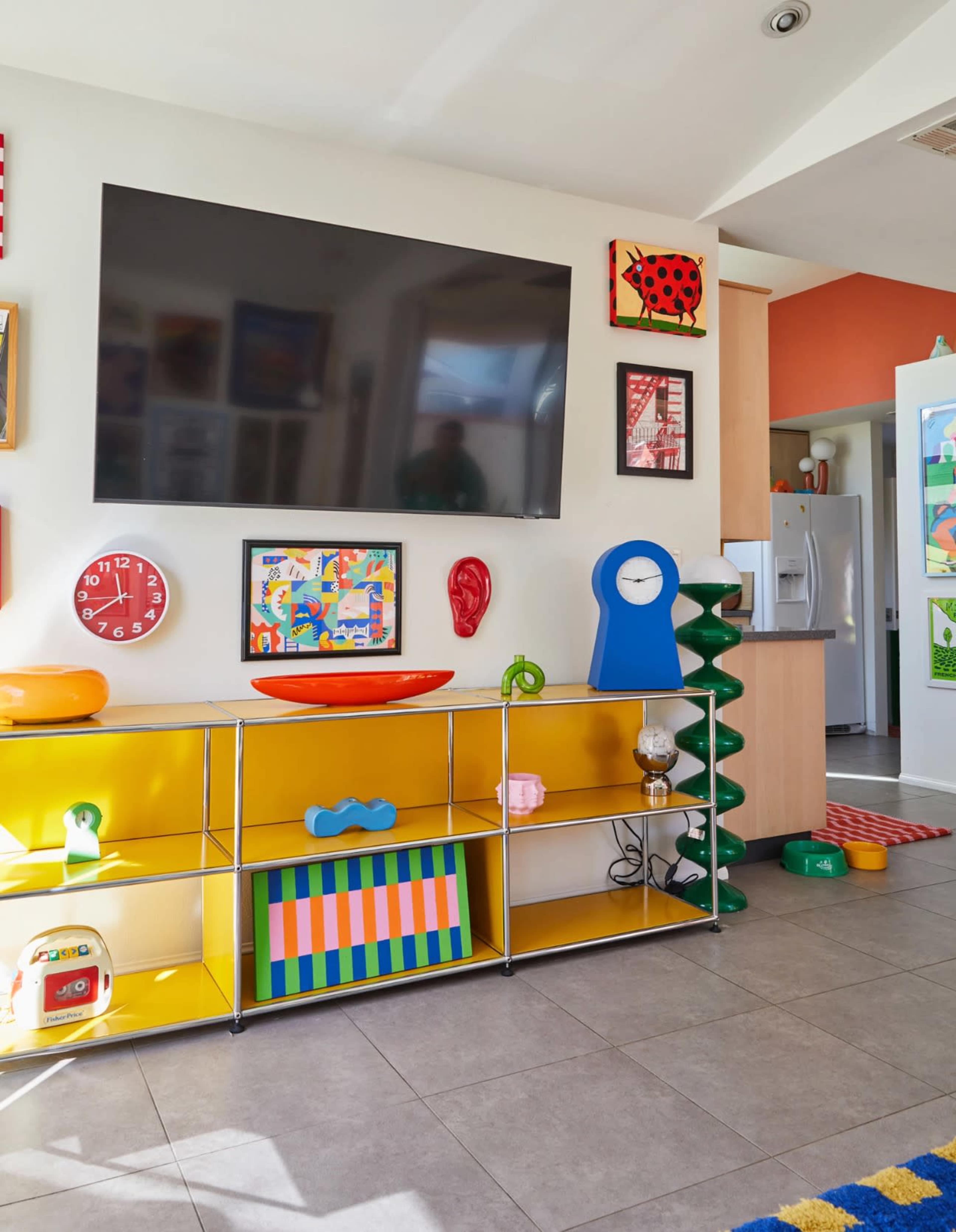 The image features a modern living room with a wall-mounted TV and colorful decorative items on a shelf, alongside framed artwork and a brightly designed rug.